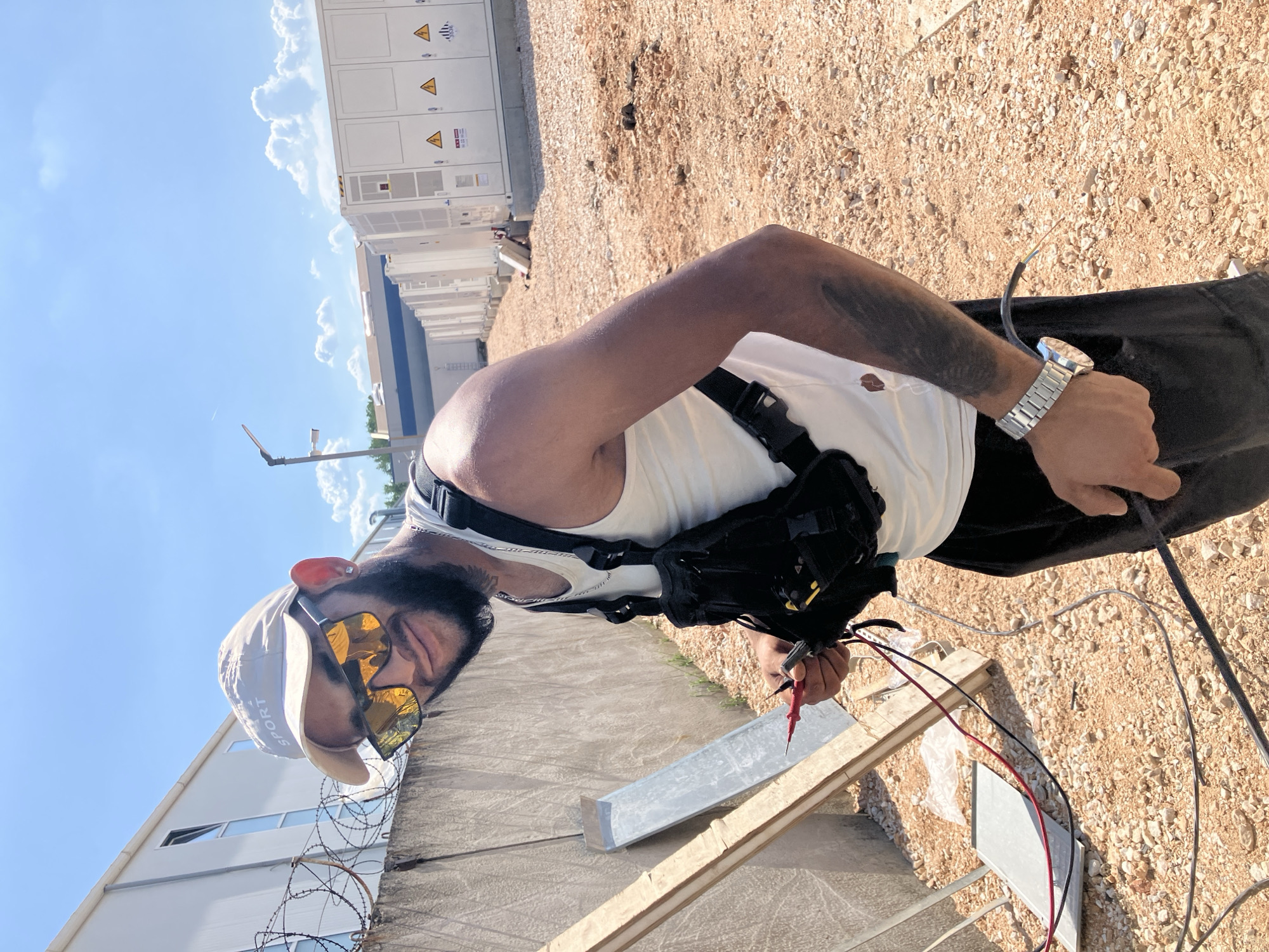 The image shows a man holding electrical probes near some exposed wires. He is wearing a white tank top, black shorts, a silver watch, and sunglasses. He also has a tool bag strapped to his chest. The location appears to be an outdoor industrial area, with buildings, gravel, and wires visible in the background. Specific to Plovdiv, Bulgaria, this image may have been taken at a site where new photovoltaic panels are installed because the city has seen a surge of installations in the recent years.