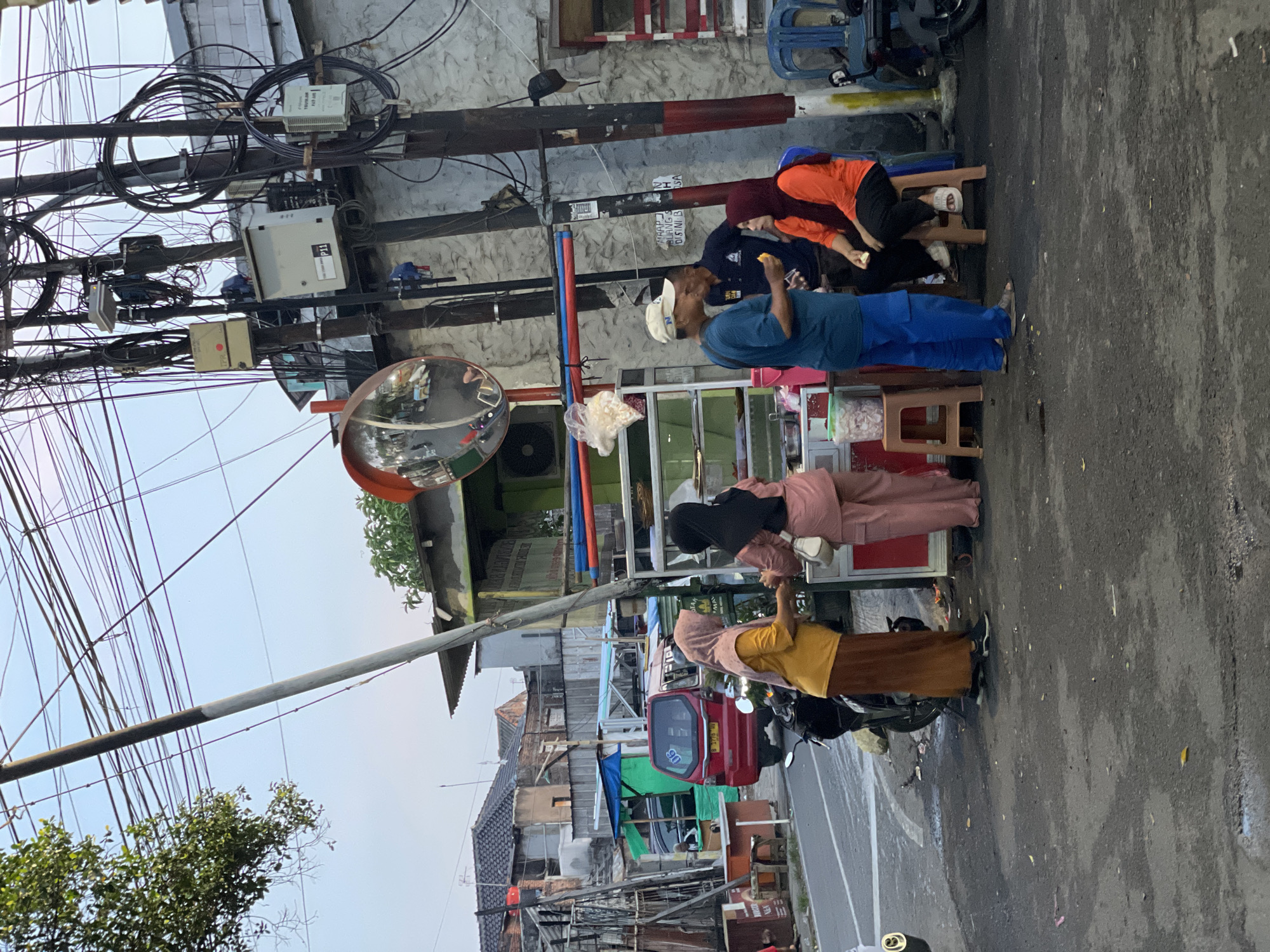 The image shows a street scene in North Jakarta, Indonesia. Four individuals are standing by a vendor stand on the side of the road. One person is wearing a blue shirt and blue pants, one wearing an orange shirt and black pants, one wearing a pink top and black head covering, and another wearing a yellow top and brown skirt. The vendor stand is silver with red trim and displays food items. A red and green three-wheeled vehicle is parked nearby. Overhead, power lines create a dense network against a light sky. A convex traffic mirror is mounted on a pole. Buildings line the opposite side of the street, showing signs of urban life.