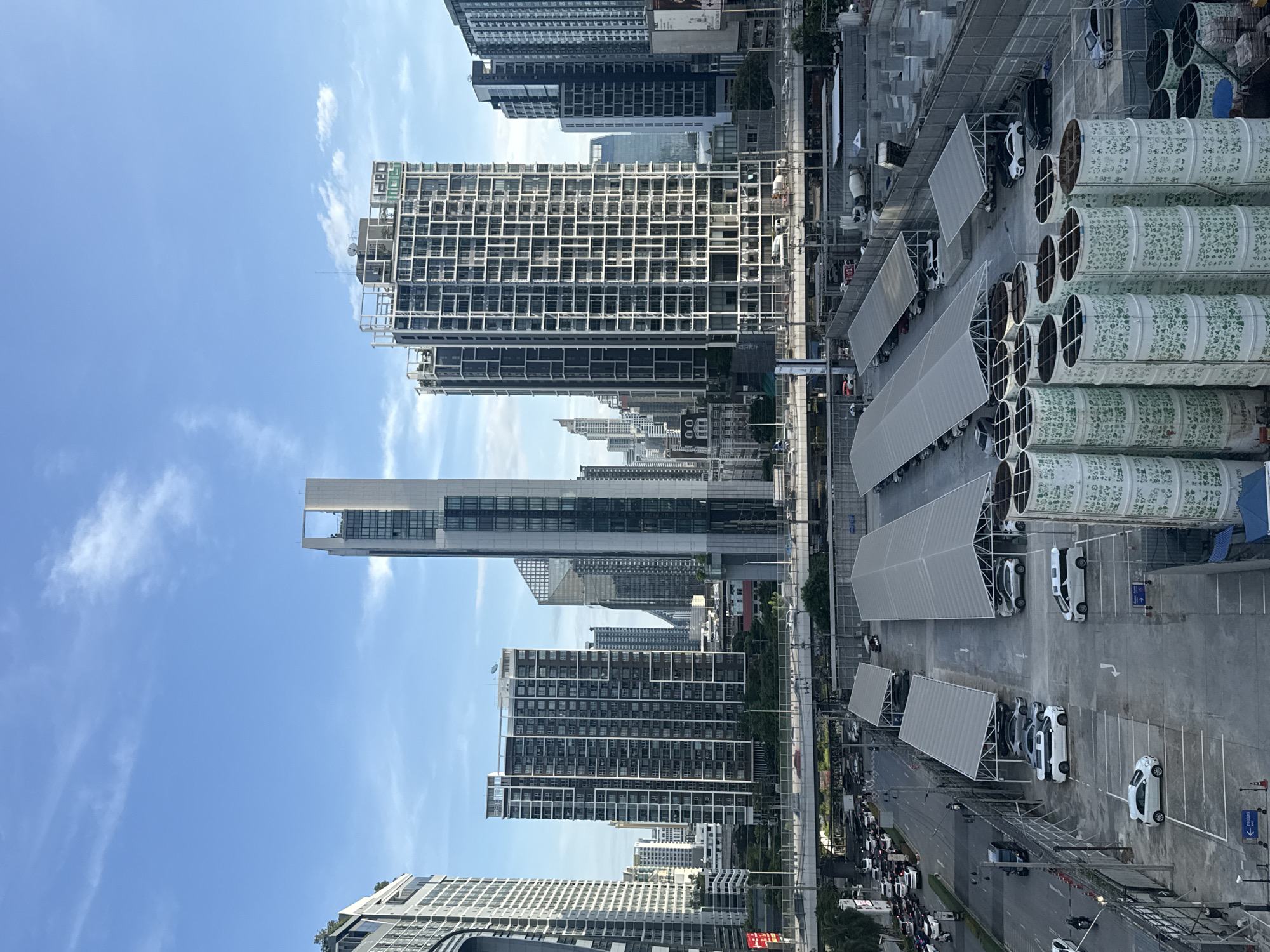 This is an eye-level, aerial shot taken from a high vantage point overlooking a busy urban scene in Bangkok, Thailand, likely during daylight hours given the bright sky and shadows. The foreground is dominated by a large, outdoor, partially covered parking lot. Numerous cars are parked in designated spots, with a mix of white and dark-colored vehicles, including sedans, SUVs, and vans. Some of the parking bays are covered by large, angled, light-colored tarps supported by metal frames. In the very foreground, to the right, are several large, cylindrical structures, stacked on top of each other, decorated with a repeating green floral pattern.
Behind the parking lot, a multi-lane road is visible, congested with traffic. Cars, motorcycles, and a few trucks are navigating the road. A raised overpass or bridge runs across the middle ground, with vehicles moving on it.
The background features a dense cityscape of modern high-rise buildings. These include office towers, apartment complexes, and other commercial structures. The architecture is varied, with many buildings characterized by glass facades and numerous windows. Notably, a distinctive tall, slender building with a rectangular cutout near its top stands out. The sky above is a clear blue with scattered wispy white clouds. The overall impression is one of a thriving, modern metropolis with ongoing urban activity.