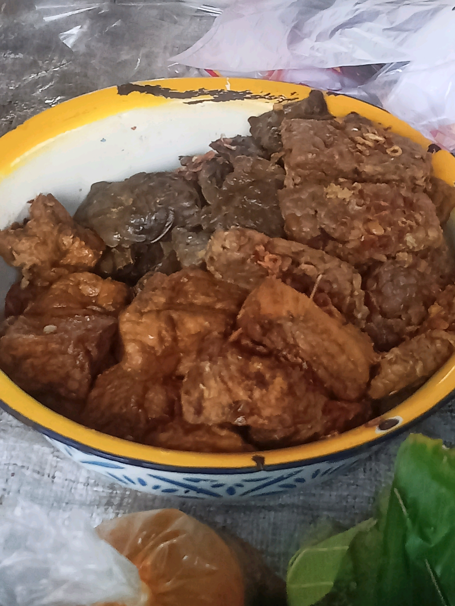 In Nganti, Indonesia, a close-up shot captures a bright yellow and white enameled bowl filled with fried foods. The bowl has a chipped rim, revealing a dark underlayer in places. The food appears to be a mixture of dark, possibly crispy pieces and lighter brown, fried morsels. In the background, out-of-focus elements suggest a market or outdoor setting, with hints of plastic bags and green leafy vegetables. The overall impression is of a casual, possibly street food presentation.