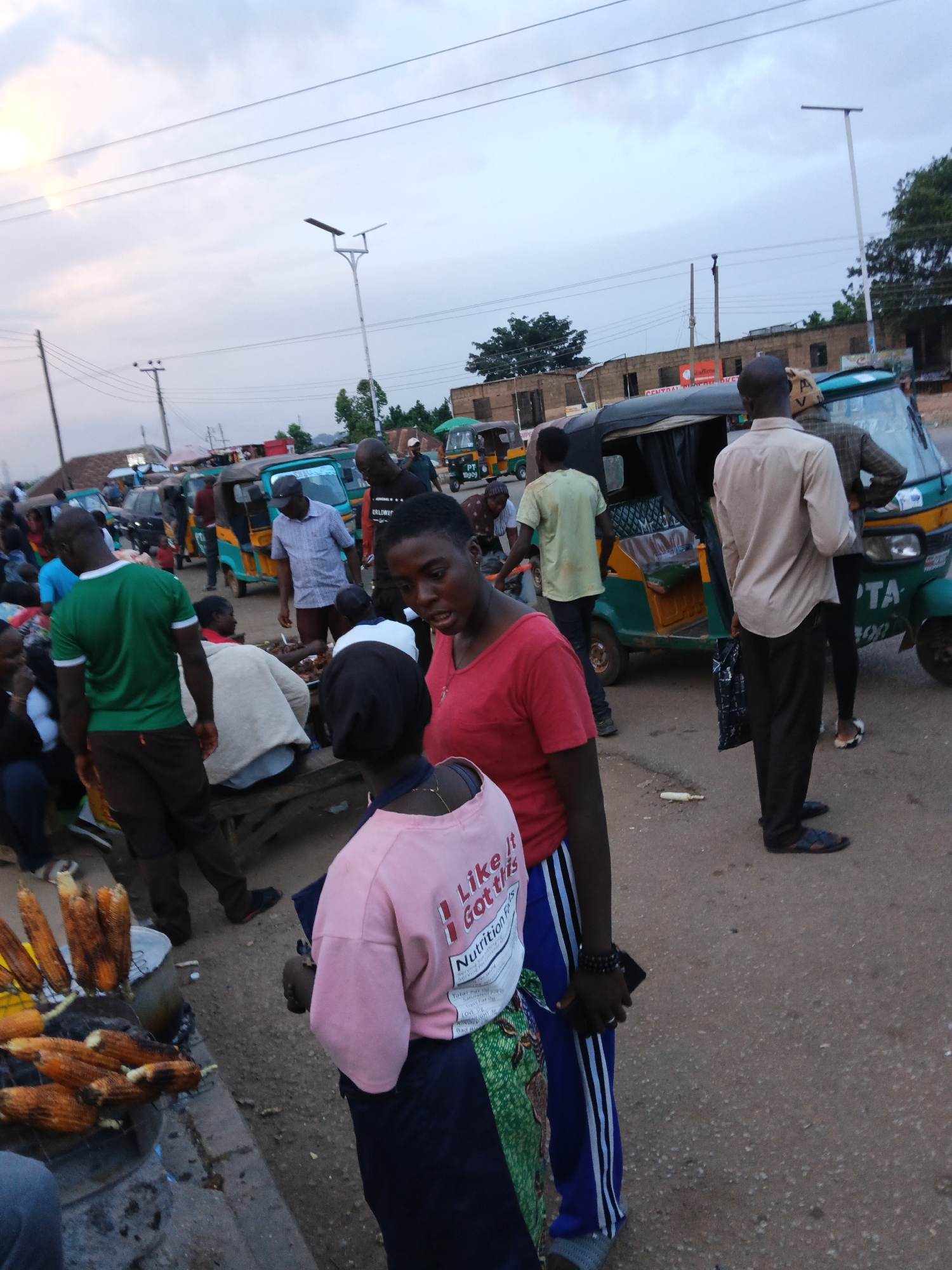 The image depicts a busy street scene in an outdoor market, possibly in a city given the presence of multiple auto-rickshaws, power lines, and buildings. The sky is overcast, suggesting it might be late afternoon or early evening. A group of people are gathered, some standing, some sitting, and others appearing to be engaged in transactions.
In the foreground, a woman in a pink t-shirt with "Like it Got This Nutrition Facts" printed on it is facing away from the camera, interacting with another person. The person she is interacting with is wearing a dark headscarf and a green and white patterned skirt. Next to them, a man in a green t-shirt and brown pants is also present.
Further back, several auto-rickshaws, common forms of transportation in many parts of the world, are parked or moving. One auto-rickshaw, painted green and yellow, has the text "PT 19909" on its rear. People are seen around these vehicles, some appearing to be passengers or drivers.
The market area features stalls with food items, including what appear to be roasted corn on the cob displayed on a metal grill. Bags and other goods are also visible. The ground is a rough, paved surface. The overall atmosphere suggests a bustling marketplace with daily activities in progress. The presence of auto-rickshaws and the architectural style of the buildings hint at a location in a developing country, possibly in Africa or Asia.