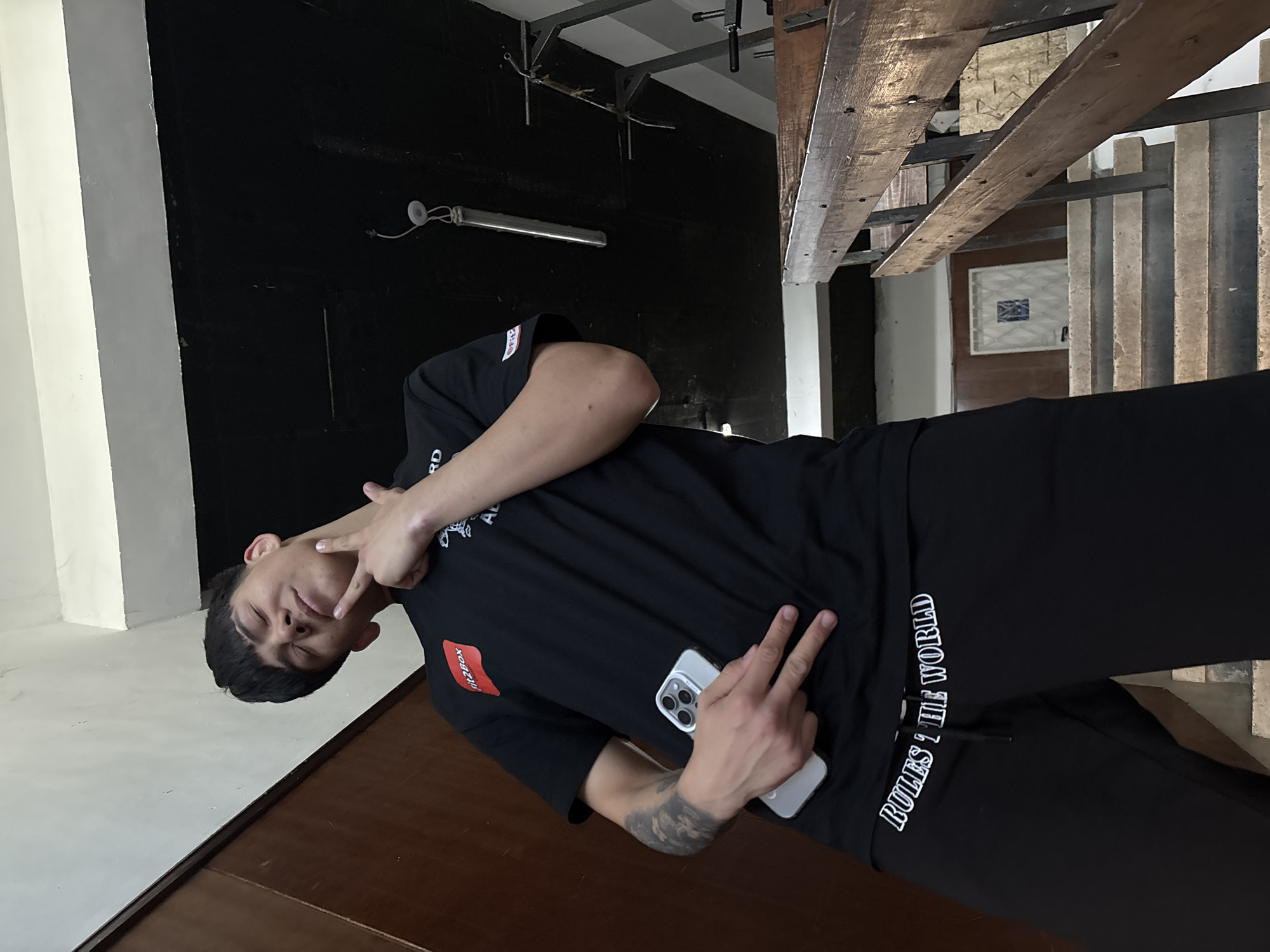 A young man is posing for a photo indoors, likely in Sofia, Bulgaria. He is positioned at an angle, looking towards the camera with a relaxed expression. He is wearing a black t-shirt and black pants, both featuring visible text. His right arm, which has a visible tattoo, is bent with his hand near his face, making a "peace" or "victory" sign. His left hand is holding a modern smartphone (appears to be an iPhone) and also making a "peace" sign.
The visible text on his clothing includes "RD AL" on his t-shirt, and a small red rectangular patch with "ritizbox" printed on it. On the side of his pants, the text "RULES 'T' THE WORLD" is clearly visible in white, stylized font.
The background suggests an industrial or contemporary interior. There's a dark wall behind him with a visible light fixture. Above and to his right, wooden beams and metal structures are part of the ceiling or a mezzanine level. Below the beams, concrete stairs or flooring are visible. The overall lighting is somewhat dim, with some brighter areas suggesting natural light might be entering from an unseen window or opening. The setting appears to be a casual environment, possibly a studio, loft, or modern living space.