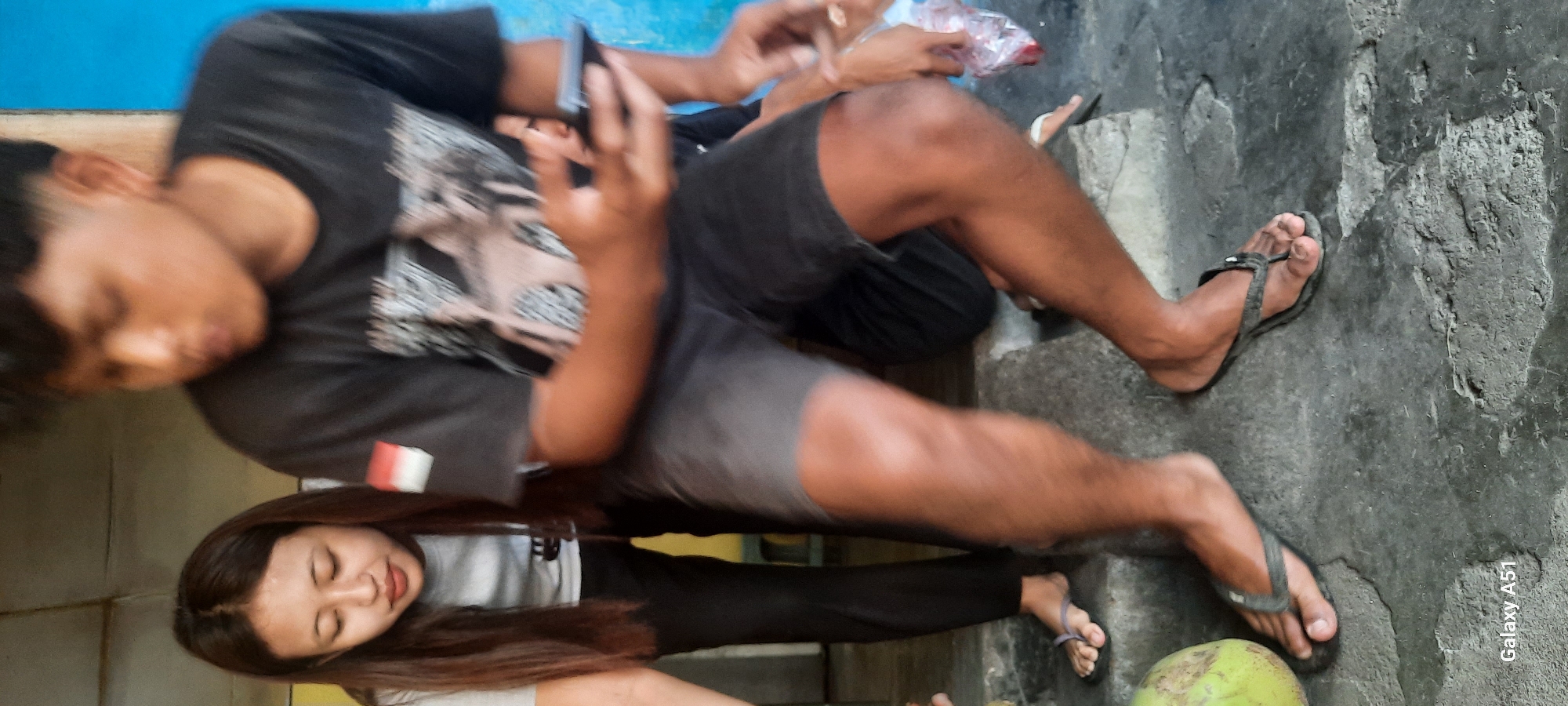 In Selopuro, Indonesia, two individuals, a male and a female, are depicted. The female, with long dark hair, is wearing a grey t-shirt and black pants, with flip-flops on her feet. The male, wearing a black t-shirt and dark shorts, is holding a mobile phone in his right hand. Both are standing on a concrete surface, with stairs visible behind them. A green coconut is situated to the left of the frame. The backdrop includes a blue wall. The image was captured with a Samsung Galaxy A51.