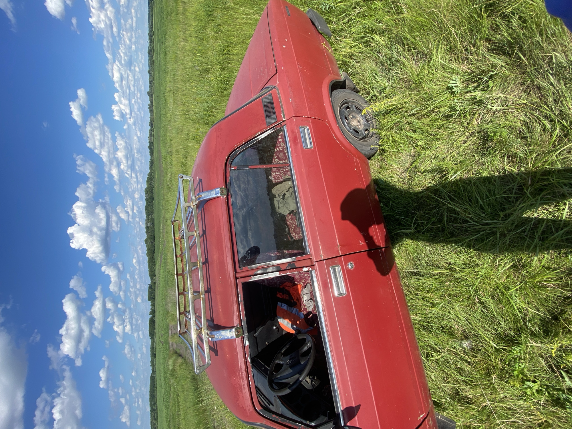 A red sedan with a chrome roof rack is parked on a dirt path in a grassy field under a blue sky with white clouds. The driver's side door of the car is open, revealing the steering wheel, an orange safety vest, and patterned upholstery. The car appears weathered and slightly dusty, with visible rust spots. The field is lush and green, with tall grass extending to the horizon. In the background, trees and a cultivated field are visible. The scene is illuminated by bright sunlight, casting a long shadow from the car and the photographer.