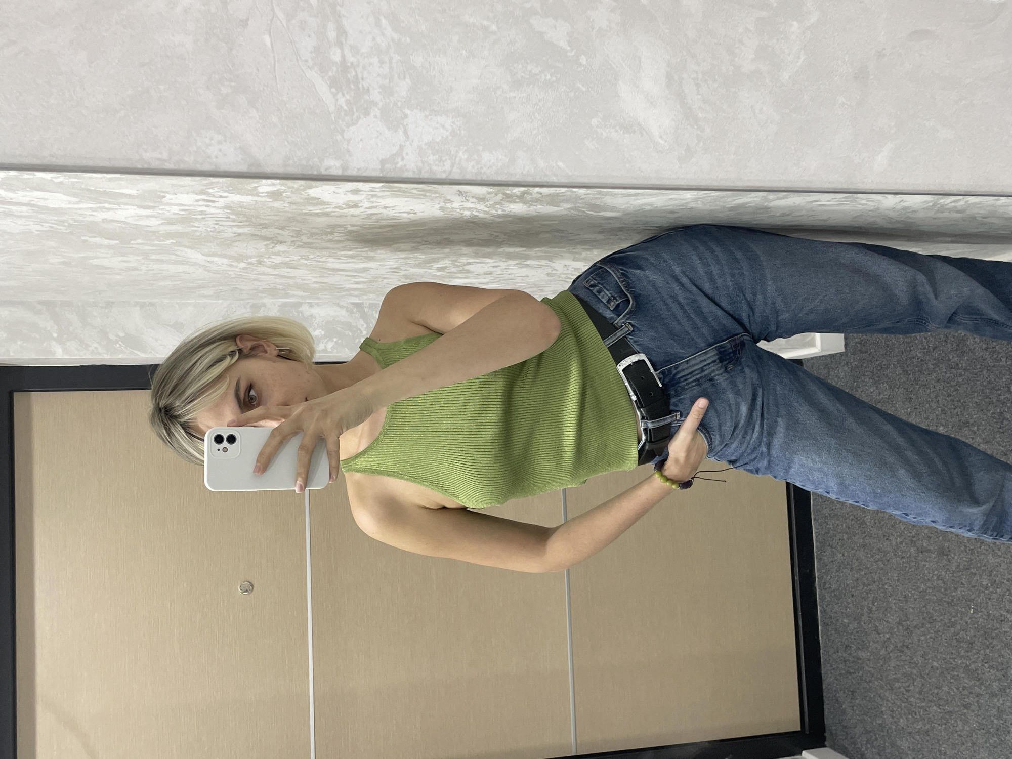The scene is an indoor mirror selfie. A female subject with short blonde hair and a visible brown eye is depicted, wearing a light green ribbed sleeveless tank top, blue jeans, and a black belt with a silver buckle. She holds a white smartphone with her left hand, which largely obscures her face. Her right hand is placed into her jeans pocket, and a green and purple beaded bracelet is visible on her right wrist. The background to the left features a light brown door with a central silver door viewer and a dark frame. To the right, a textured light gray wall is visible. The floor is covered with dark gray carpet. The specific location within Izhevsk, Russia, cannot be determined from the available visual information.