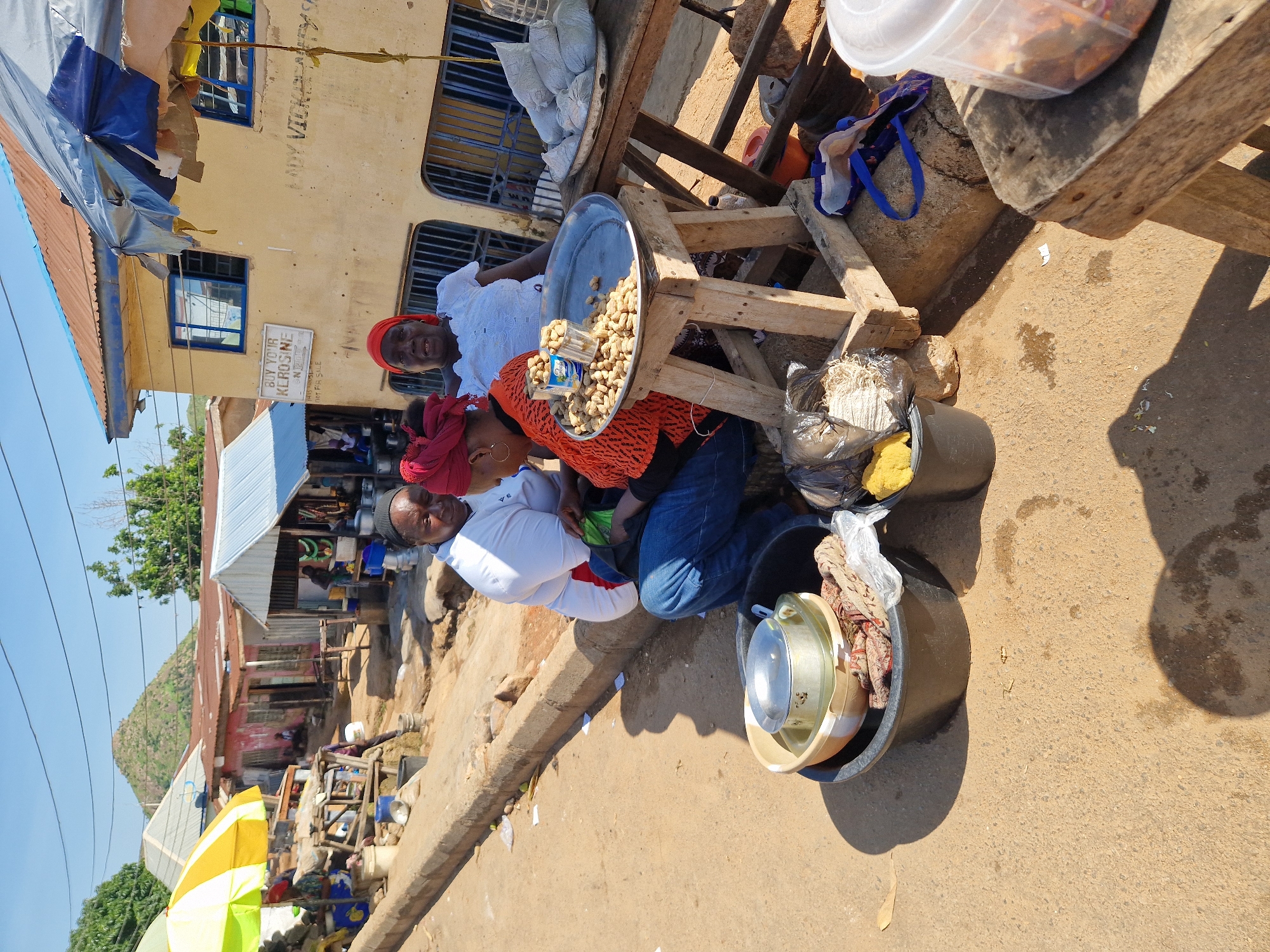 The image depicts an outdoor market scene with three women engaged in commerce. Two women are seated, while one stands behind them. The seated women are interacting, with one holding a green bag and the other examining nuts displayed on a metal tray. The standing woman, wearing a white top and red headscarf, appears to be a vendor. Various items are visible, including buckets, metal pots, packaged goods, and a wheelbarrow. The environment suggests a dry, dusty location with a building in the background that has windows and a sign reading "BUY YOUR KEROSINE". The overall scene is a candid snapshot of everyday life in a market setting.