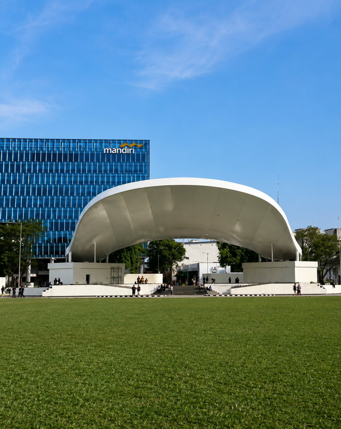 In a city park in Medan, Indonesia, a wide green lawn stretches towards a modern outdoor amphitheater. The amphitheater features a white, curved roof supported by white walls, with tiered seating in front of it. Behind the amphitheater, a tall, blue glass building with horizontal white lines displays the orange "mandiri" logo on its upper section. Several people are scattered across the lawn and seated on the amphitheater's steps. The sky is a clear, bright blue with a few wispy clouds. Trees are visible on either side of the amphitheater, and in the background, other buildings and structures can be seen.