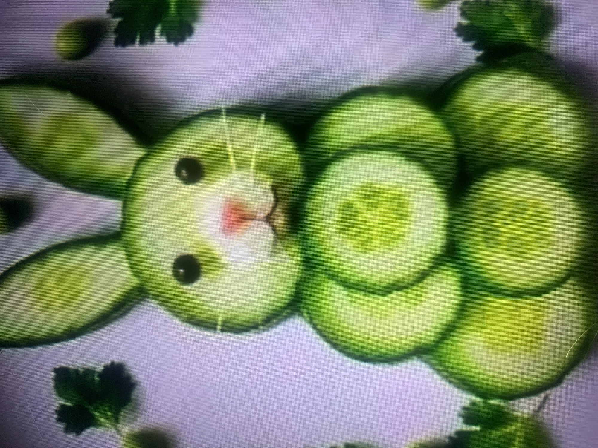 The image displays a close-up, overhead shot of a rabbit crafted from cucumber slices and other vegetables, arranged on a white surface. The rabbit's ears are elongated cucumber slices. Its head is a round slice of cucumber, featuring two black olive pieces for eyes, a pink radish slice for a nose, and white cucumber slivers for whiskers. The body is constructed from several stacked, round cucumber slices, showing the inner seeds. The rabbit is flanked by sprigs of parsley and small green spheres, possibly olives, on either side, and a few more are scattered behind the rabbit's head. The background is a plain, light purple-tinted surface, likely a tablecloth or countertop. The lighting is bright, casting subtle shadows. There are no people or discernible urban elements visible in the image, and therefore, no specific location within Almaty, Kazakhstan, can be determined.
