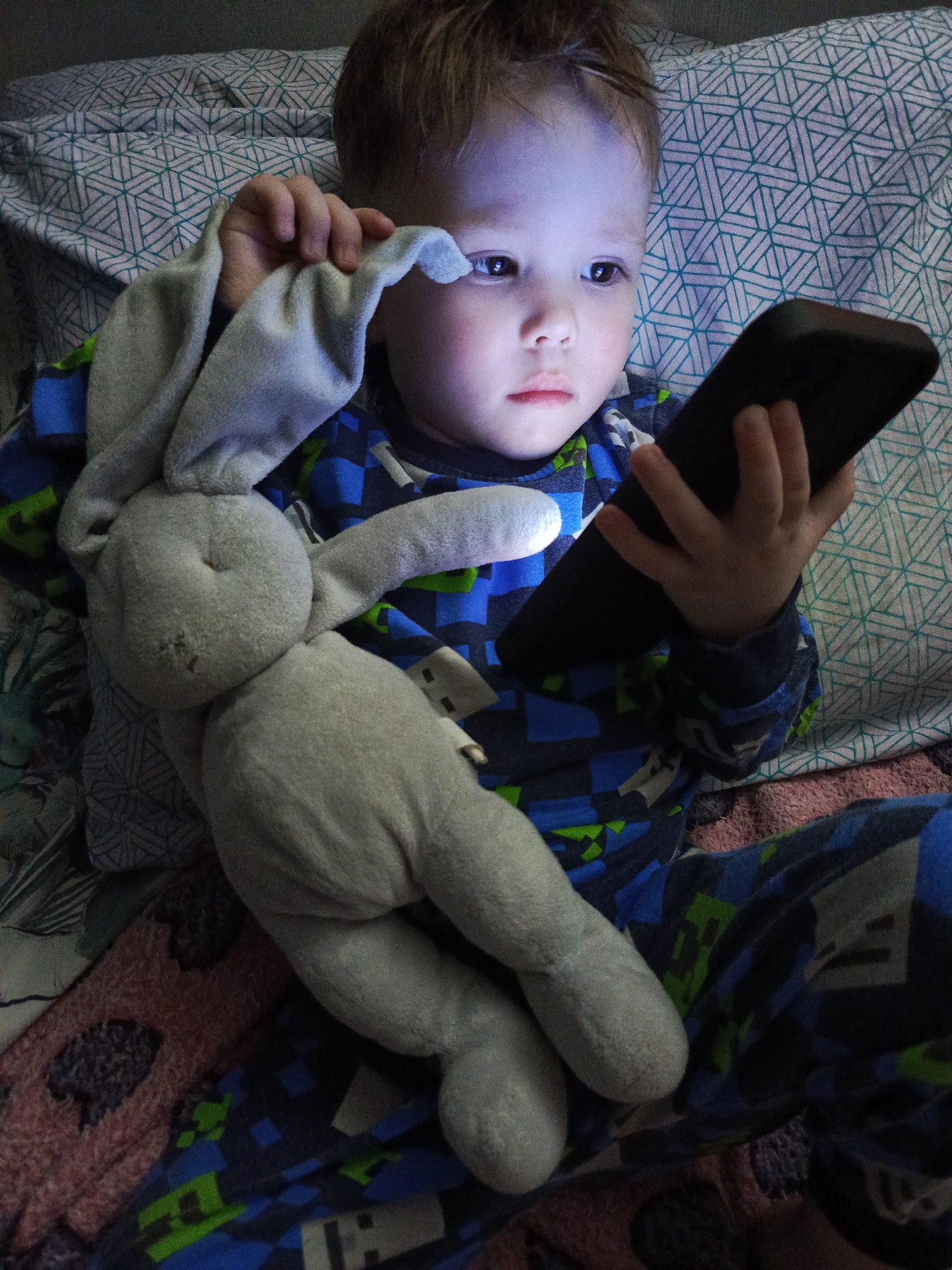A young child, approximately 3-4 years old, lies in bed in Chaikovsky, Russia. He is wearing blue pajamas with a geometric pattern. The child holds a black smartphone in his right hand and has a gray stuffed bunny resting on his chest. He uses his left hand to partially cover his eye with the bunny's ear. The screen of the smartphone illuminates his face. The bedding consists of a pillow with a geometric pattern and a blanket with a blurred design.