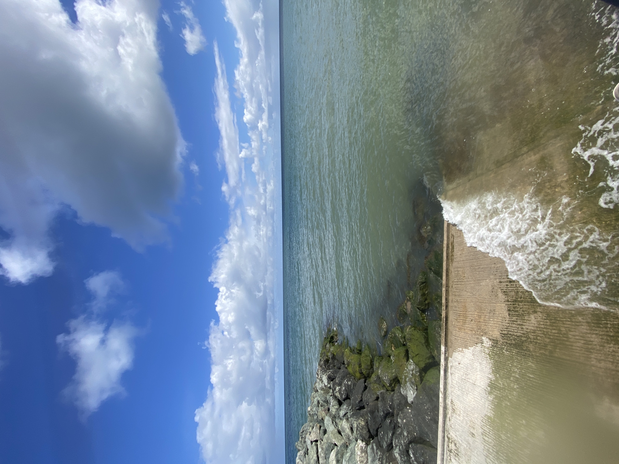 The photograph, taken in Old Colwyn, United Kingdom, shows a concrete embankment abutting the sea. The sky, predominantly blue, has fluffy, white clouds dominating the upper left portion of the image. The ocean surface is a murky green-brown and slightly choppy. Waves break against the embankment. Rocks with green algae line the bank.