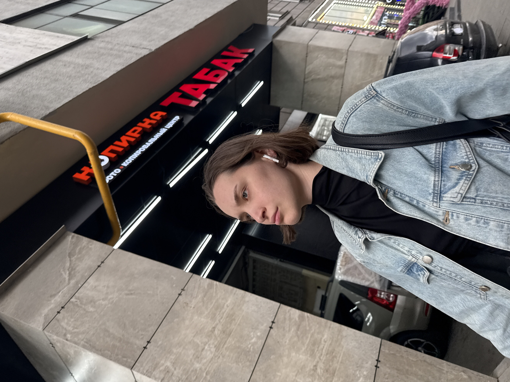 A young woman with light brown hair, wearing a light blue denim jacket over a black turtleneck and white wireless earbuds, stands outside in Moscow, Russia. She is looking to her left. Behind her, a building features a large red illuminated sign with Cyrillic lettering that reads "НА МИРКА ТАБАК" and smaller text below it. A yellow pipe runs diagonally above the sign. A light-colored SUV is visible through an open garage door to the left. To the right, a black car is parked on the street, with a building facade featuring glowing lights in the background. The woman is carrying a black shoulder bag. The overall environment suggests an urban setting.