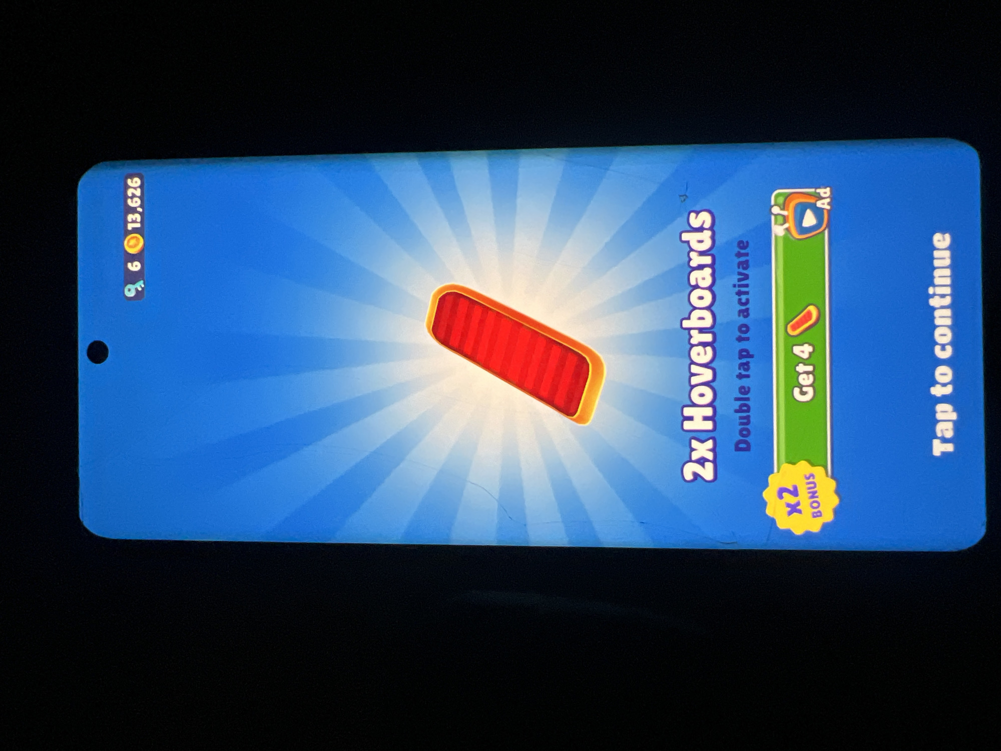 The image displays a mobile game interface, likely from "Subway Surfers," on a blue background with radiating lines. In the center, a red hoverboard with an orange glow is prominently featured. Above it, the text "2x Hoverboards" appears in purple, followed by "Double tap to activate" in lighter blue. To the left of the "Double tap to activate" text is a yellow starburst graphic with "x2 BONUS" written inside. Below this, a green banner with rounded ends contains the text "Get 4" and an icon of a red hoverboard. To the right of the "Get 4" text and hoverboard icon is an orange television-like icon with a play button, labeled "Ad." At the bottom of the screen, the text "Tap to continue" is displayed in white. In the top right corner, the game displays "6" followed by a key icon, a coin icon, and the number "13,626." The image quality is somewhat poor with visible scratches on the screen. There are no discernible elements that would allow for the localization of this image to Lagos, Nigeria.