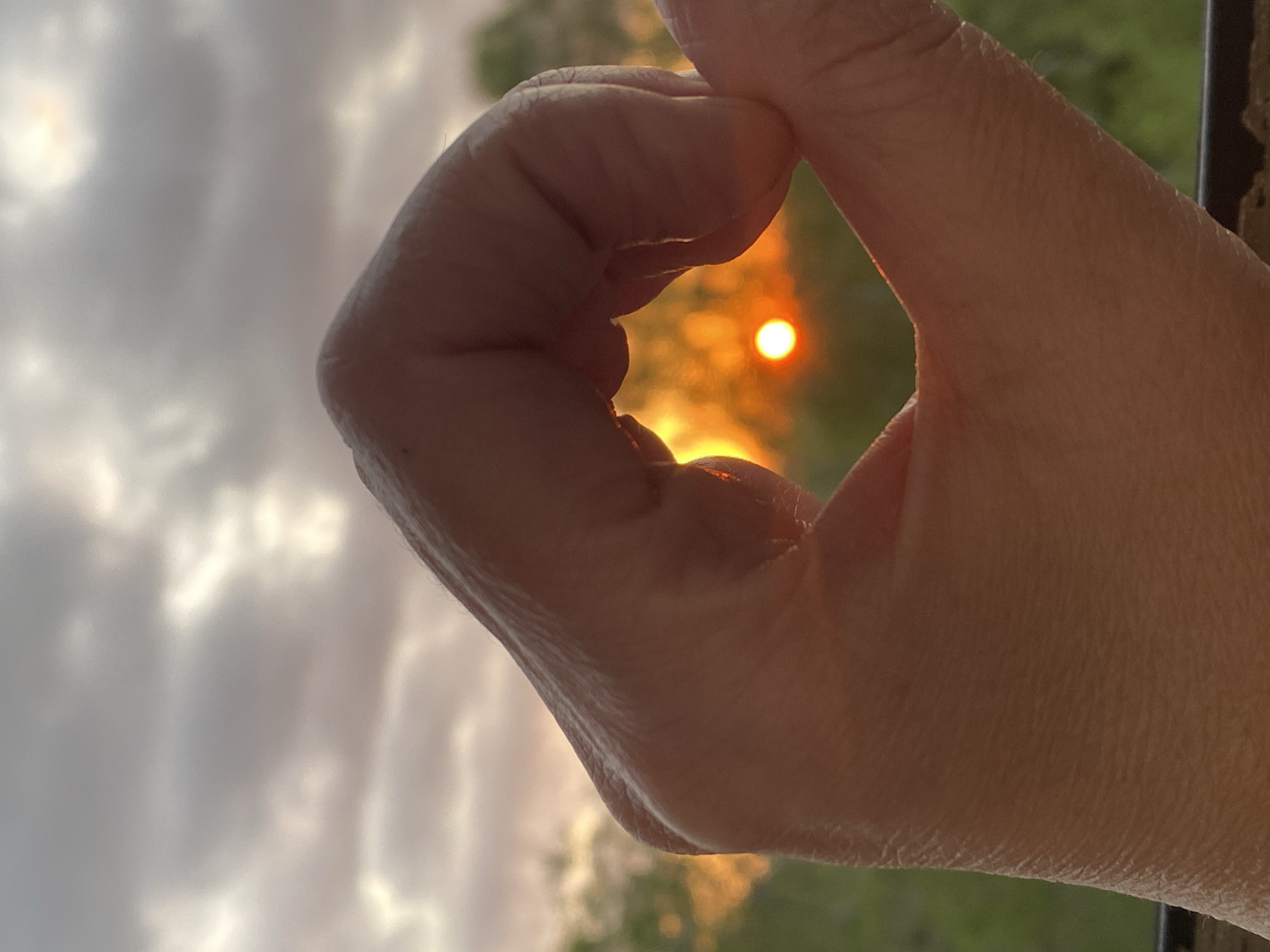 A human hand forms an "OK" sign against a backdrop of a sunset with the sun visible through the circle made by the hand. The background shows a scene of water and sky, possibly a body of water reflecting the sunset. Details of the surrounding environment are obscured by the focus on the hand and the setting sun. The location is not discernible from the image.