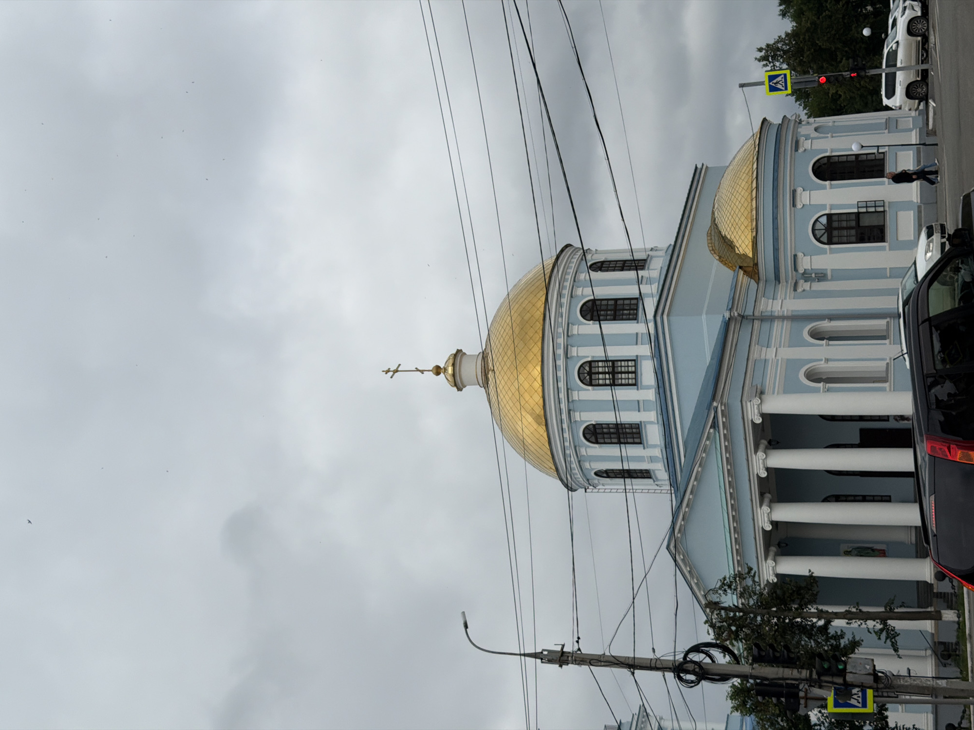 The image shows the Resurrection Cathedral in Votkinsk, Russia, on a cloudy day. The light blue building features a prominent golden dome topped with a cross, and a portico with white columns. Overhead, a network of power lines crisscrosses the sky, partially obscuring the building and the overcast sky, which contains several small, indistinct dark specks, possibly birds or debris. To the left, a street light and traffic signals are visible. A pedestrian crossing sign with a blue figure is also present, along with a red traffic light. In the foreground, the back of a black SUV is visible, and to its right, a white SUV and a person walking on the sidewalk. The environment suggests an urban setting.