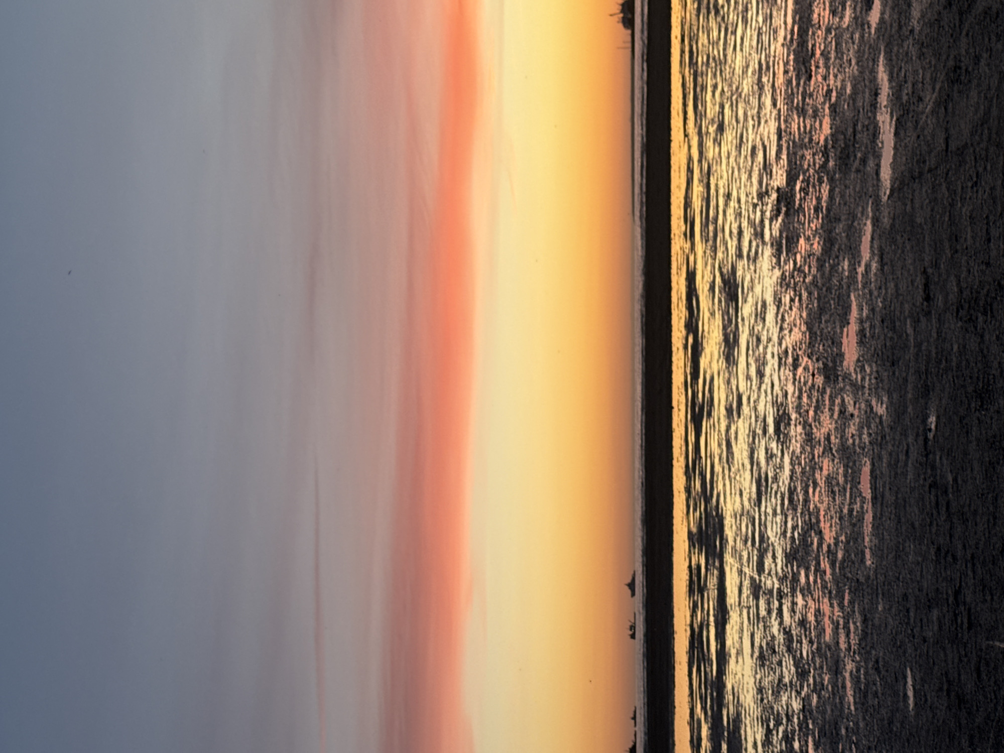 The image depicts a serene seascape at dusk, likely from the coast of Tallinn, Estonia. The sky is a gradient of soft blues and pinks, transitioning into vibrant oranges and yellows near the horizon, indicative of a sunset. Several silhouetted ships are visible in the distance, appearing as small dark shapes against the colorful sky. The foreground features a dark, wet, and reflective surface, possibly a beach or mudflats, with pools of water mirroring the sky's colors. The overall atmosphere is tranquil and visually striking, capturing the beauty of a coastal twilight.