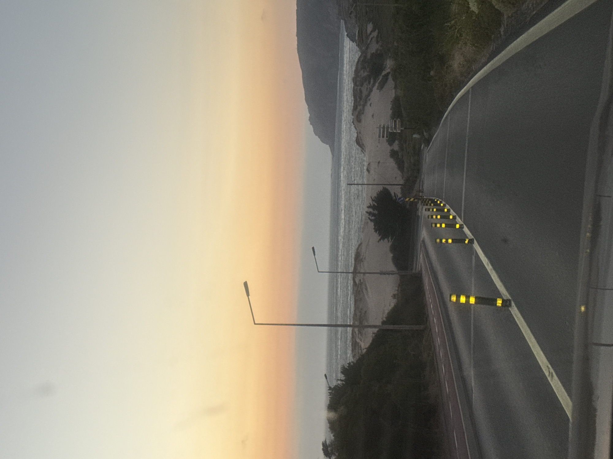 The image depicts a scenic view of a coastal road at dusk, likely near Cascais, Portugal. The road, paved with asphalt, curves gently to the right, with a yellow line marking its edge. Along the left side of the road, a series of black bollards with yellow reflective bands are spaced out, delineating the lane. A separate bike lane, marked with a red surface and white lines, is visible to the left of the main road.
In the background, sand dunes rise up, covered with sparse vegetation. Beyond the dunes, the ocean stretches out, its surface reflecting the soft hues of the sunset. The sky transitions from a pale blue at the top to warm shades of orange and yellow near the horizon. A distant, rocky coastline is visible on the right side of the frame. Two streetlights are present, one taller and closer, the other shorter and further away, both silhouetted against the twilight sky. The overall atmosphere is tranquil and serene, capturing the beauty of a coastal sunset.