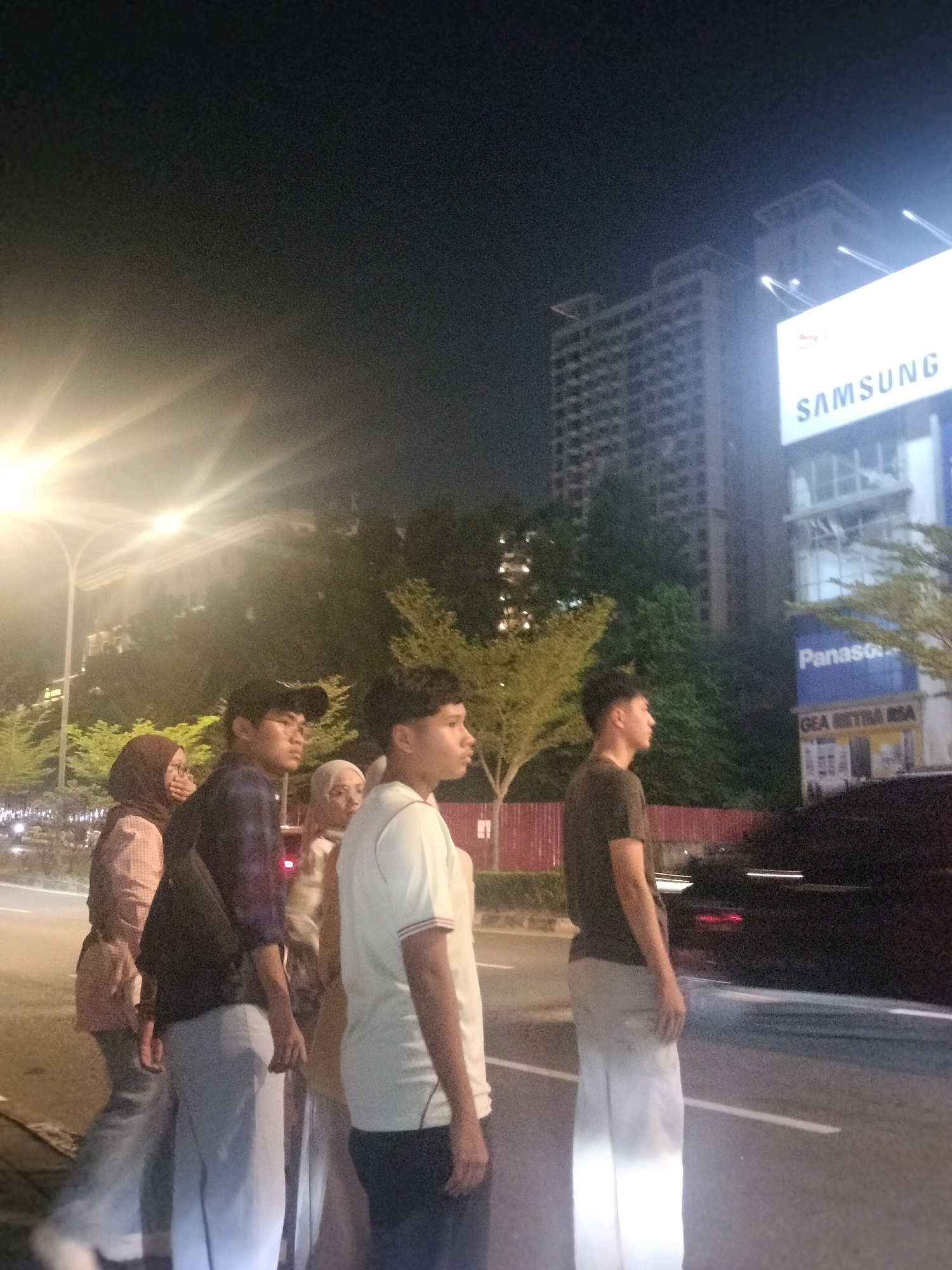 In Batam, Indonesia, at night, a group of five individuals stands near a street. From left to right, a woman in a pink patterned shirt and headscarf faces away from the camera. Next to her, a man wearing glasses and a dark shirt with a backpack is partially visible. A third person, also wearing glasses and a light-colored headscarf, stands behind them. A young man in a white t-shirt with red trim and dark pants faces forward. The person furthest to the right is a young man in a dark t-shirt and light-colored pants. In the background, there are trees, streetlights, and two tall buildings. One building prominently features a large "SAMSUNG" sign, and below it, a "Panasonic" sign is visible, along with other store signs. A dark-colored car is parked on the right side of the street. The scene appears to be illuminated by artificial lighting, including streetlights and the illuminated signs.
