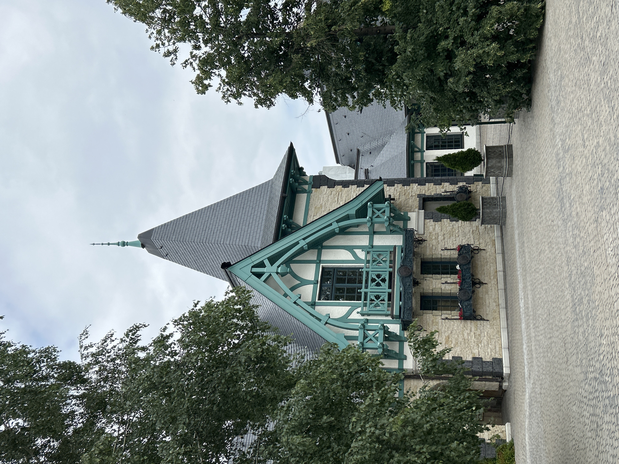 This is an eye-level, medium outdoor shot of a building in Khryashchevka, Russia. The building has a distinctive architectural style with a light-colored stone facade and white walls, trimmed with teal-colored decorative elements. A prominent feature is a steeply pitched grey tiled roof with a conical spire topped with a finial. A small balcony with a teal railing and decorative X-shaped accents is visible on the upper floor, beneath a decorative gable. The building is surrounded by mature trees with lush green foliage, which partially obscure the structure. In front of the building, there is a paved courtyard made of grey interlocking stones. Two rectangular stone planters, each containing a small, conical evergreen tree, are placed in the courtyard. The sky is overcast with a light grey hue. The scene appears tranquil and well-maintained.