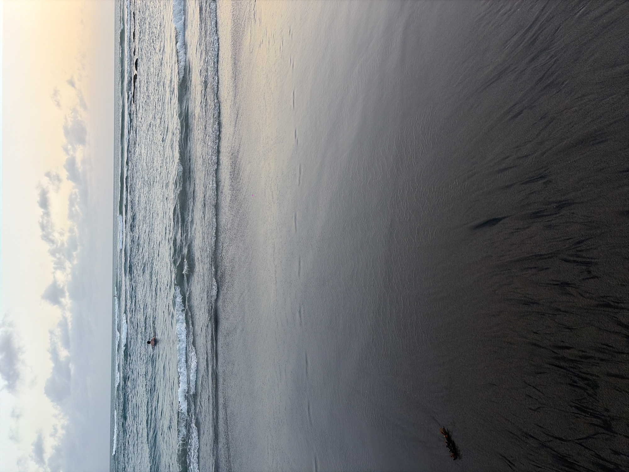 A vertical, eye-level medium shot captures a serene beach scene at sunset in North Kuta, Indonesia. The foreground is dominated by dark, wet sand, marked with gentle ripples and streaks of lighter sand, suggesting the receding tide. A single piece of dark brown seaweed rests on the sand in the lower left quadrant. The middle ground features the ocean with several waves rolling towards the shore, their white crests contrasting with the darker water. In the distance, a lone individual is partially submerged in the ocean, appearing to swim or float. The horizon line is clearly defined, separating the water from the sky. The sky itself is a pale gradient, transitioning from a soft orange hue near the horizon to a muted blue with scattered, wispy clouds towards the top of the frame. The overall atmosphere is calm and tranquil.