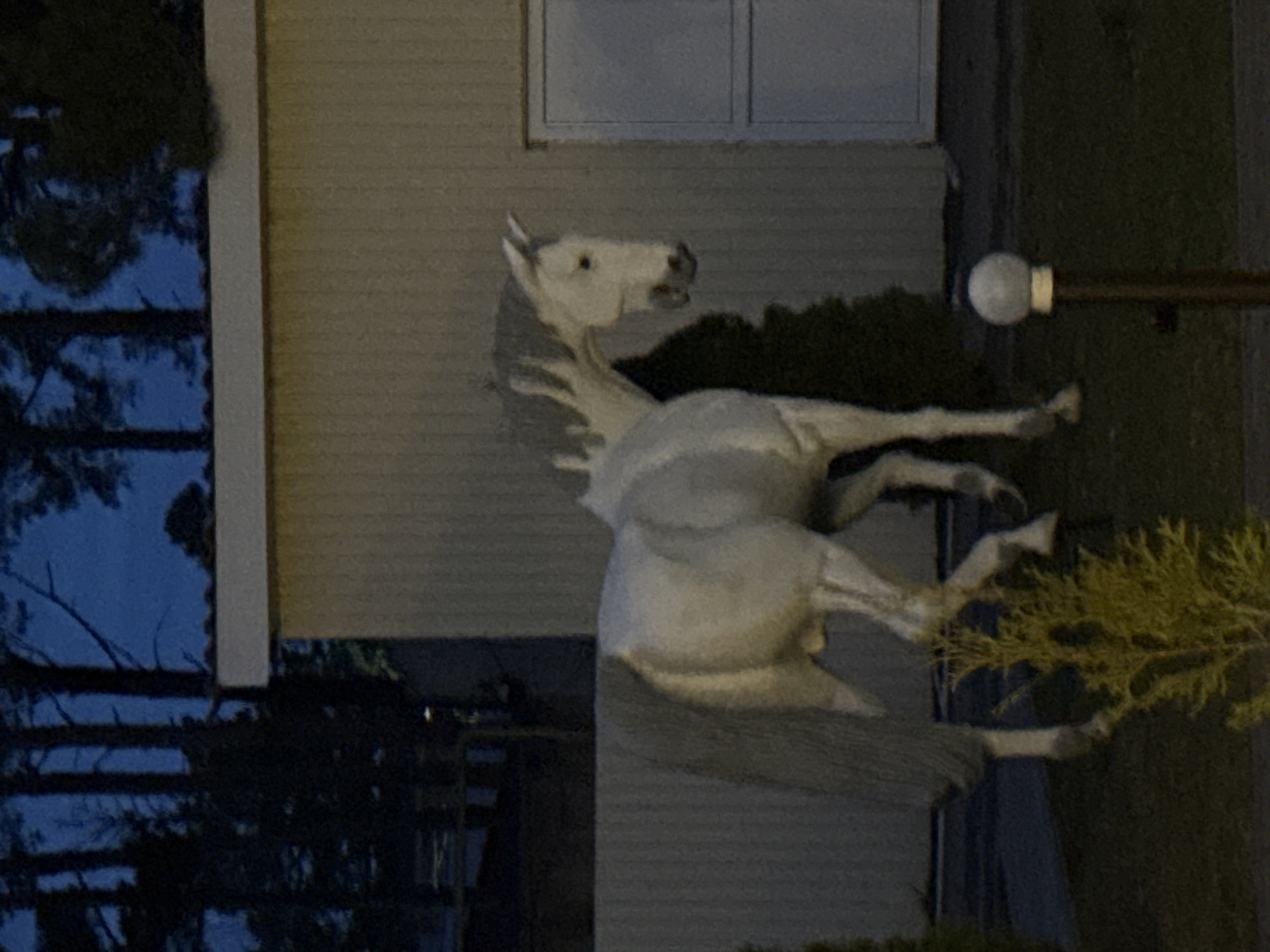 A sideways view shows a white horse statue in Votkinsk, Russia, positioned near a light pole and shrubbery. The statue is located near a window. The sky is visible through the trees on the left side.