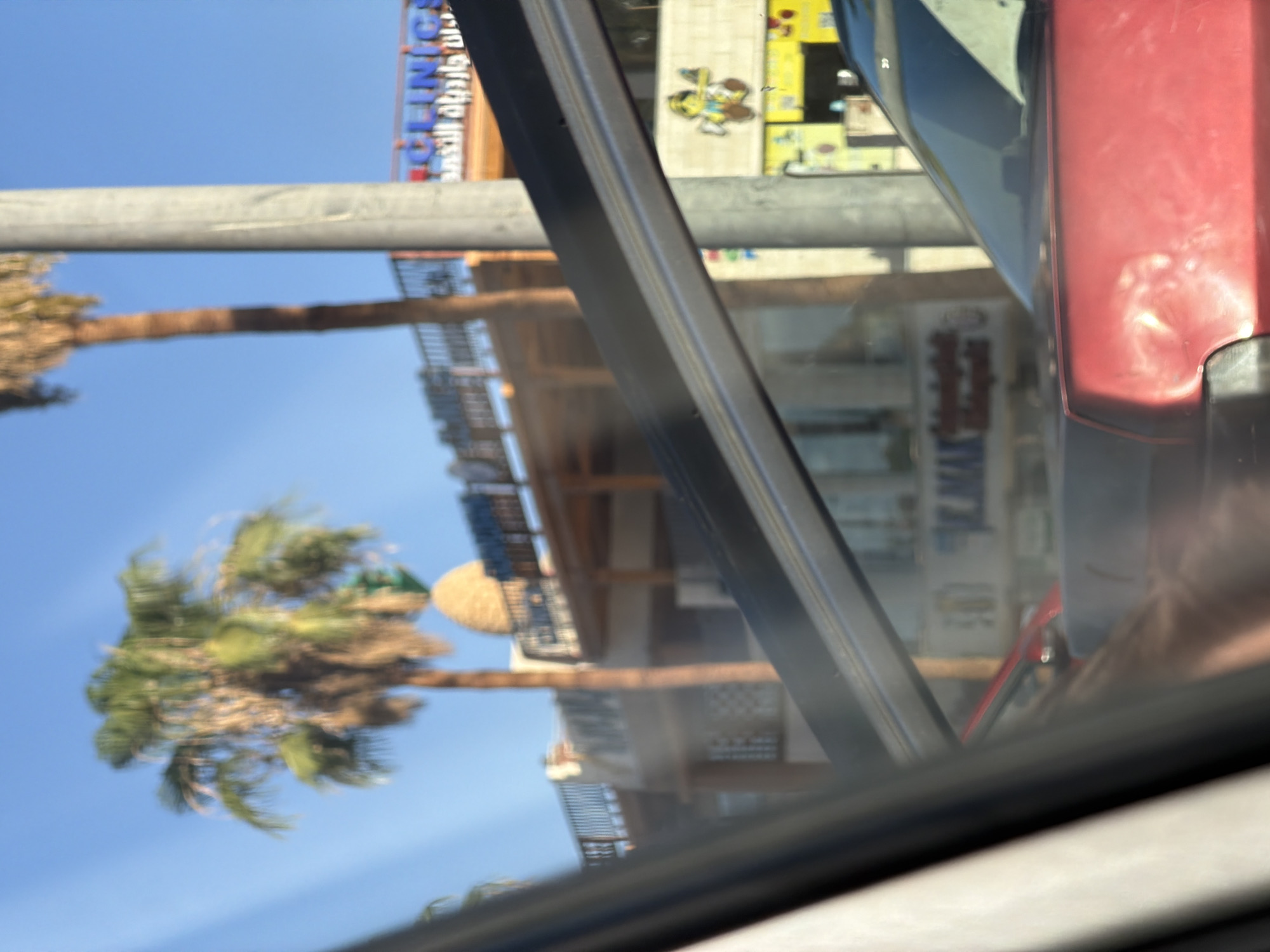 The image depicts a scene in Sharm El Sheikh, Egypt, viewed from inside a vehicle. The foreground is dominated by the interior of the car, with a black car frame and a reflection of the photographer visible on the red car's hood. In the mid-ground, a building with a balcony and awnings is visible, with palm trees in front of it. The background features a clear blue sky, with the upper portions of palm trees visible. There is also a sign with Arabic writing and the word "CLINICS" visible. A street light pole is also present. The scene appears to be a commercial area with shops and businesses. The overall impression is a sunny day in a coastal city.