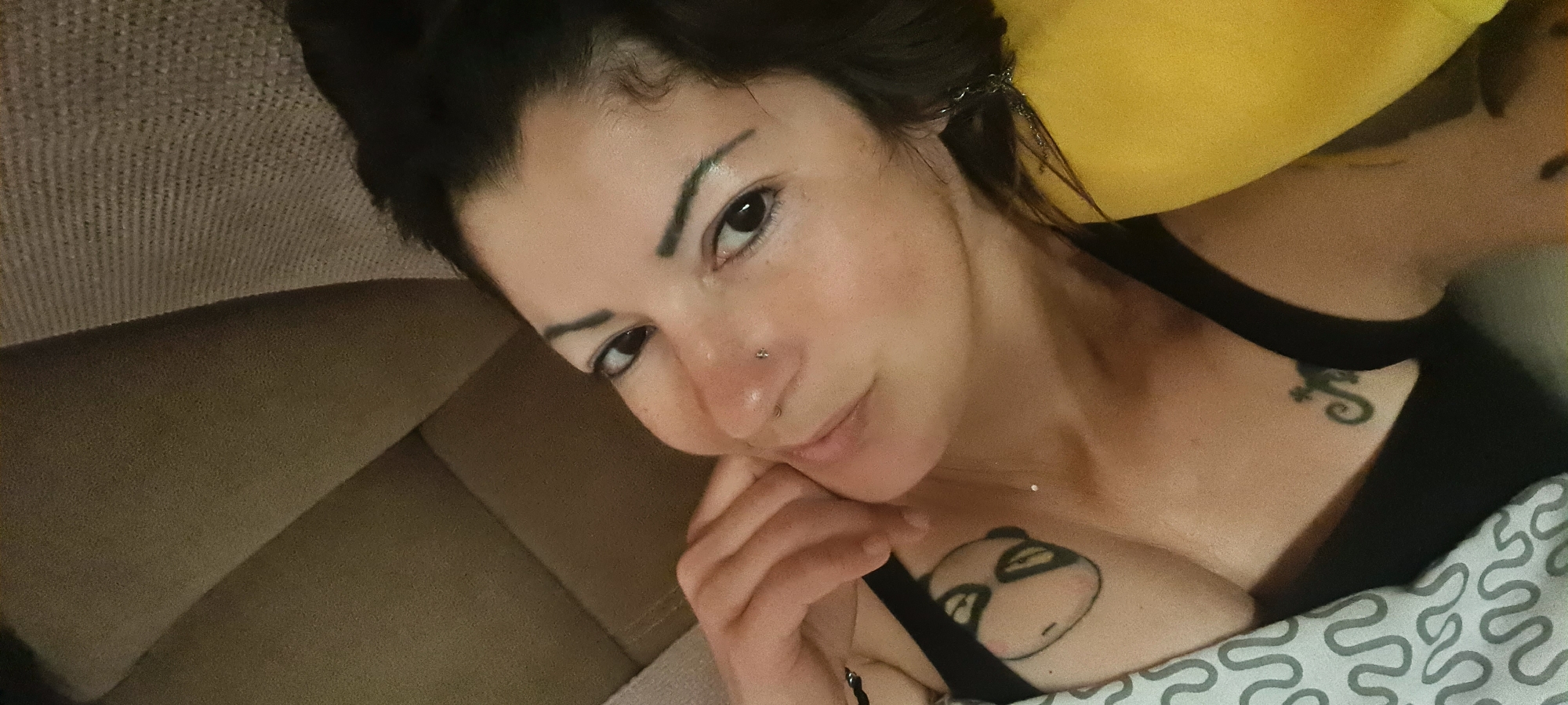 A person with dark hair is lying on a couch with a yellow pillow behind their head. They are wearing a black tank top, and a patterned blanket covers part of their chest. The person has a nose piercing and tattoos on their chest and collarbone. The background shows part of the couch and the pillow, suggesting an indoor setting. There are no identifiable clues to indicate this photo was taken in Nahariyya, Israel.