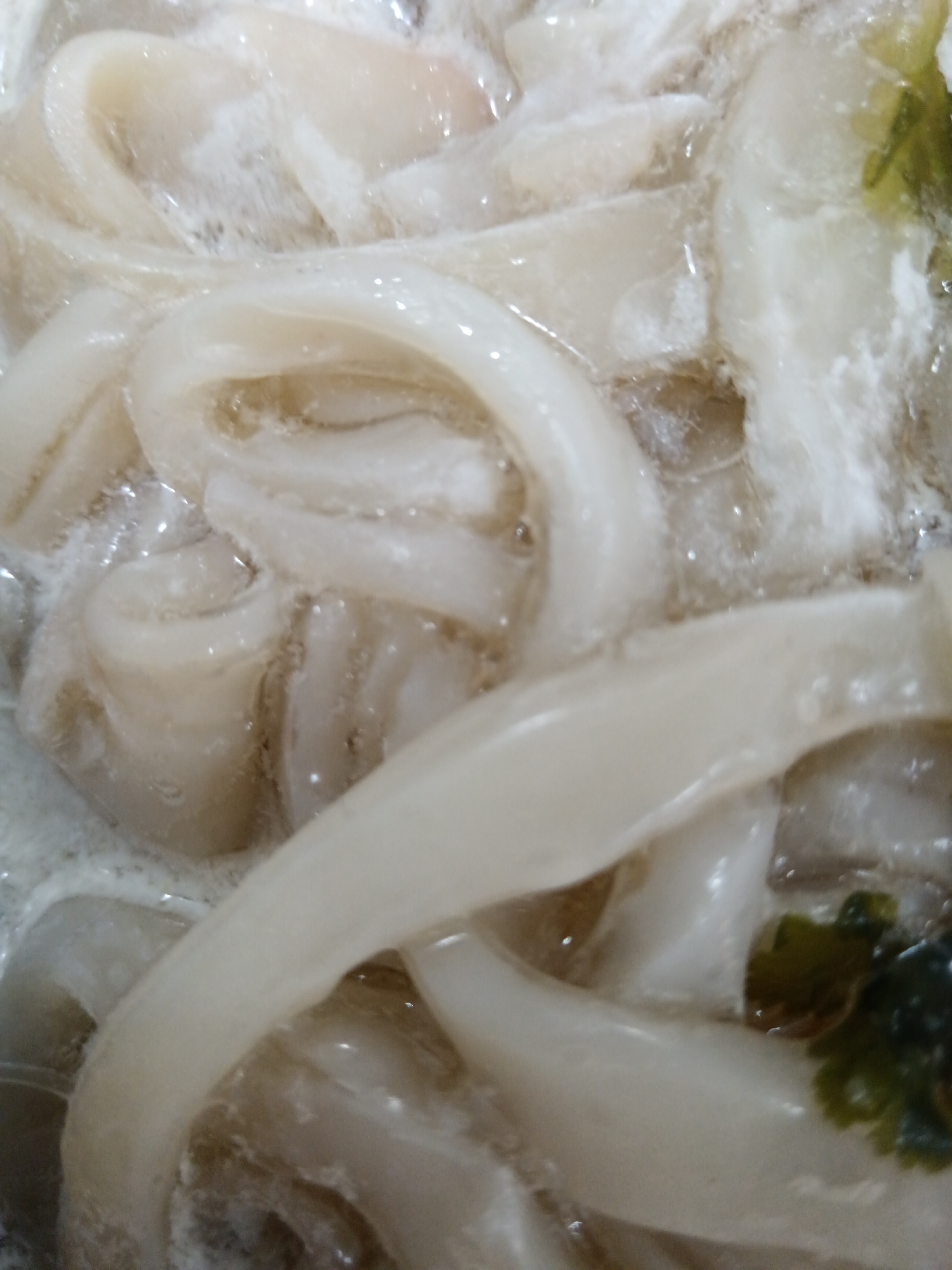 The image is a close-up, high-angle shot of uncooked squid rings in what appears to be a bowl or container, partially submerged in liquid. The squid rings are pale white and have a translucent, slightly iridescent quality. They are arranged in a coiled, overlapping fashion, creating a visually dense composition. Some areas show more foamy or bubbly texture in the liquid, suggesting it might be a brine or marinade. In the upper right and lower right corners, there are hints of green, leafy material, possibly herbs or garnishes. The lighting is diffused, and the overall impression is one of fresh, raw seafood preparation. The location in Sorochinsk, Russia, is not discernible from the image itself.