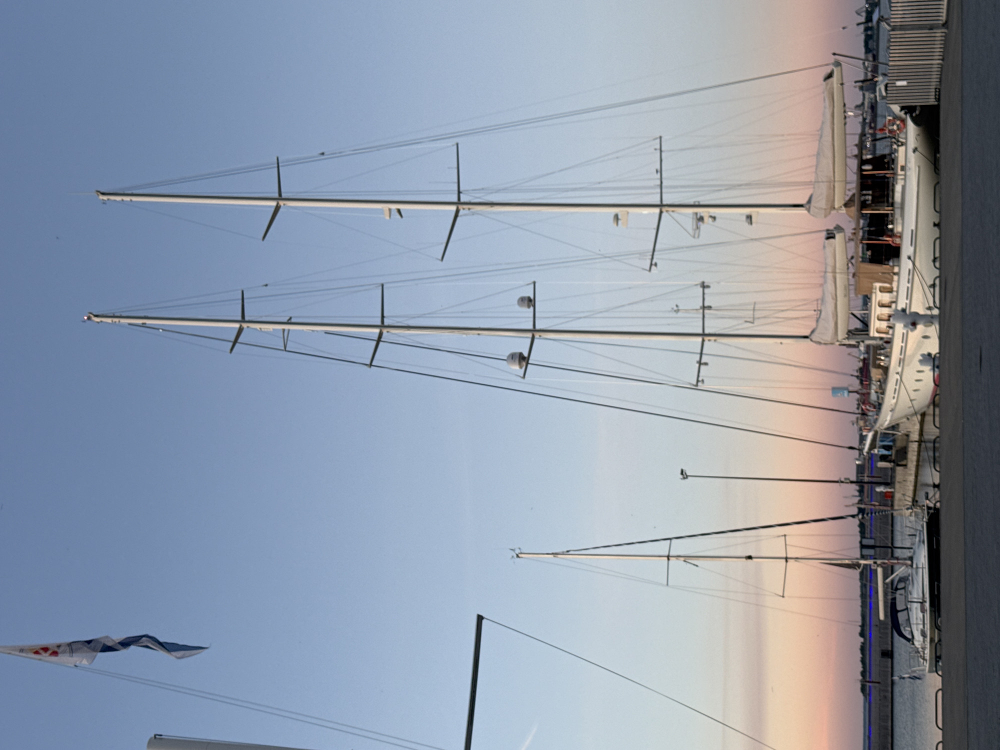 The image depicts a harbor scene at dusk in Tallinn, Estonia. The sky transitions from a clear blue at the top to soft pink and orange hues near the horizon, indicating either sunrise or sunset. Several sailboats are docked. The most prominent are two large sailing yachts with multiple masts and rigging. In the foreground, a smaller white sailboat with a dark hull is moored closer to the dock. The dock itself is made of grey concrete, and a metal fence runs along its edge. In the background, industrial elements of the harbor and a distant city skyline are visible. A flag with blue and white patterns is flying from a pole on the smaller sailboat. The overall impression is one of peaceful maritime activity during twilight.