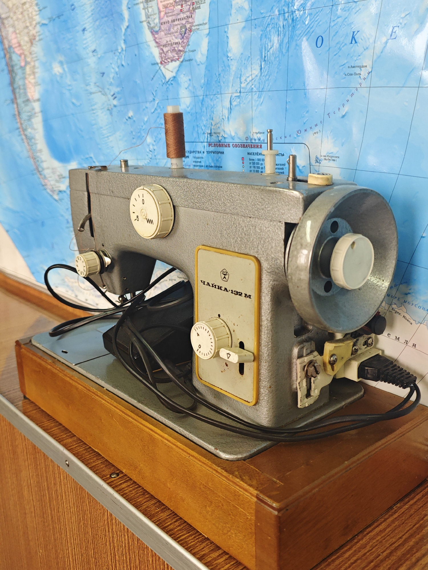 An old, gray metal sewing machine, model "Chaika-132 M", with a wooden base, is positioned on a wooden surface in front of a map of the world. The sewing machine has several dials and a spool of brown thread on top. Wires are plugged into the machine. The map in the background displays blue oceans and landmasses with place names in Cyrillic script, including "Krasnoyarsk, Russia". The lighting is natural, coming from the side. The scene appears to be indoors, possibly in a home or a workshop.