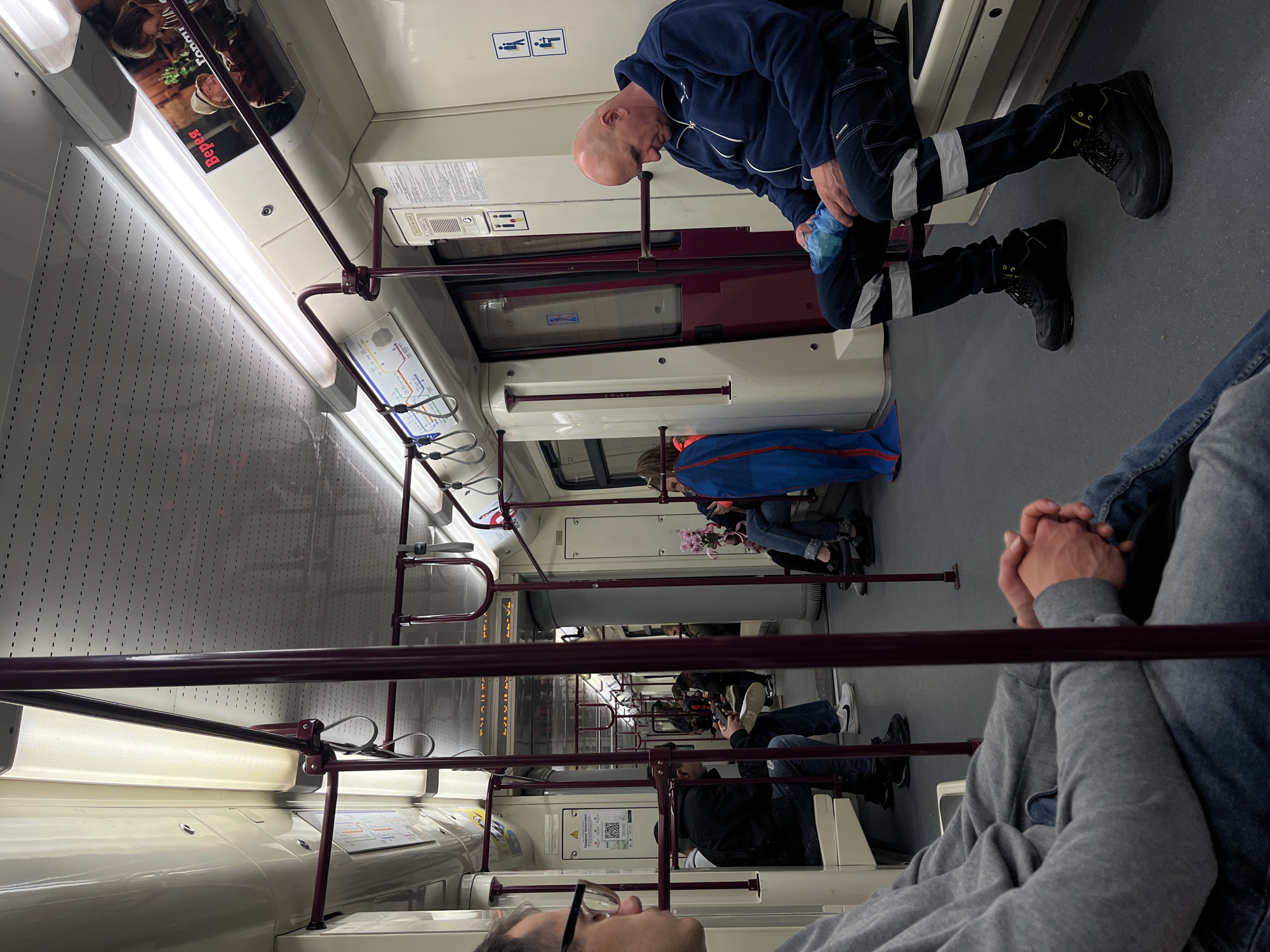 The image captures the interior of a public transport vehicle, likely a metro train or tram, in Sofia, Bulgaria. The perspective is from a seated passenger looking down the carriage. The vehicle features light-colored walls, a grey floor, and reddish-brown handrails and support poles. Overhead fluorescent lights illuminate the space.
In the immediate foreground, the head (wearing glasses) and clasped hands of a person in a grey hoodie and blue denim are visible. Further inside the carriage, a bald man, dressed in a dark blue zip-up jacket and dark work pants with reflective grey stripes, is seated or leaning against a partition. He holds a light blue plastic bag in his hands. Behind him, other passengers are visible; one person wears a bright blue jacket, and another is seen holding a bouquet of pink flowers.
The carriage walls display various information panels and route maps. A pictogram for disabled seating is visible on a panel above the man in work clothes. An advertisement on the ceiling features an image of people and the Cyrillic word "БЕРЕГ" (BEREG). Another panel clearly shows the words "Транспортна схема" (Transport scheme) and "София" (Sofia), confirming the location. A QR code is also visible on one of the information displays. The scene depicts typical passenger activity during a commute.