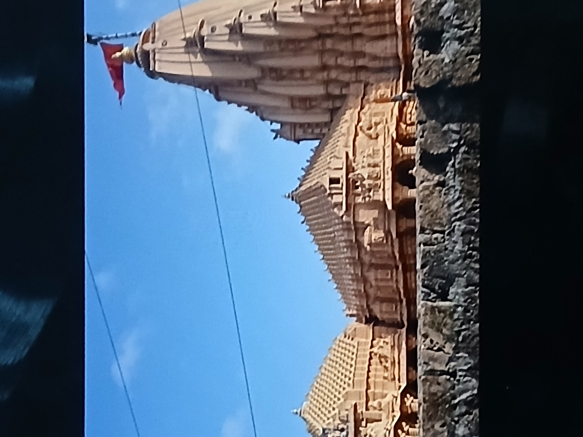 The image shows a temple structure, possibly of Hindu origin, with intricate carvings and multiple tiers, set against a clear blue sky. A red flag is visible on the topmost structure. The temple appears to be built from sandstone or a similar type of stone. A portion of a rough stone or brick wall is visible on the right side of the image. Power lines are visible against the blue sky. The temple's location is unknown. The city of Bengaluru, India, is not discernible in the image.