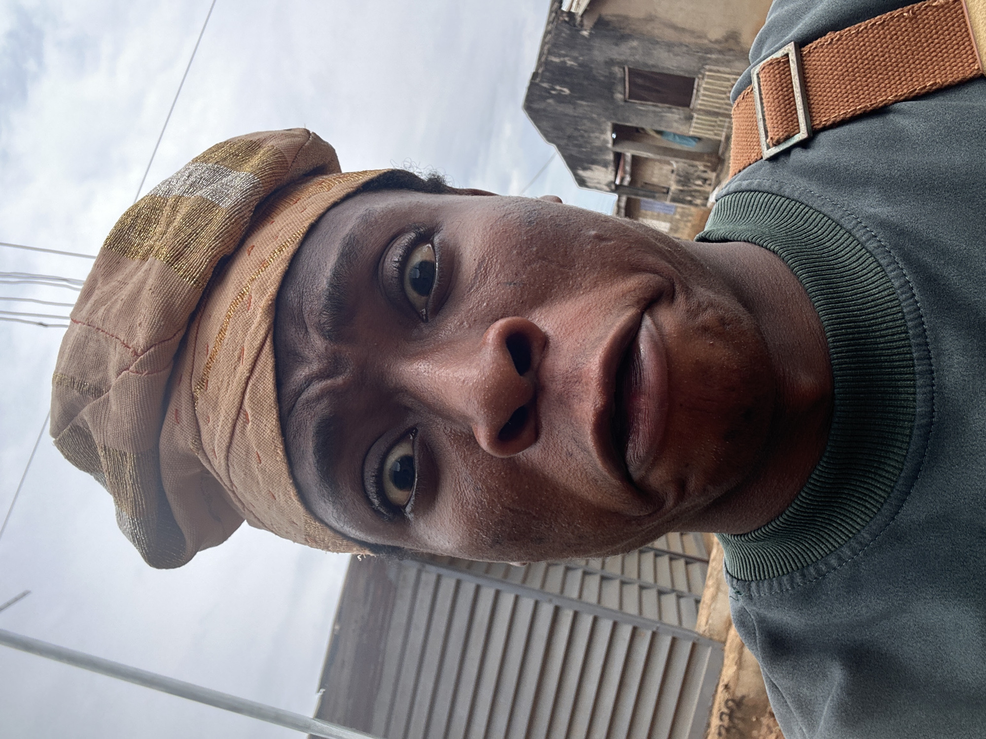 A close-up selfie depicts a dark-skinned individual wearing a brown cap with gold and silver accents. They have a slightly furrowed brow and a neutral expression. The person is wearing a dark green sweater with a brown strap or belt. The background includes a weathered building and a gray, cloudy sky. A metal gate or fence is partially visible in the lower portion of the frame. The location is in the city of Unknown, Unknown.