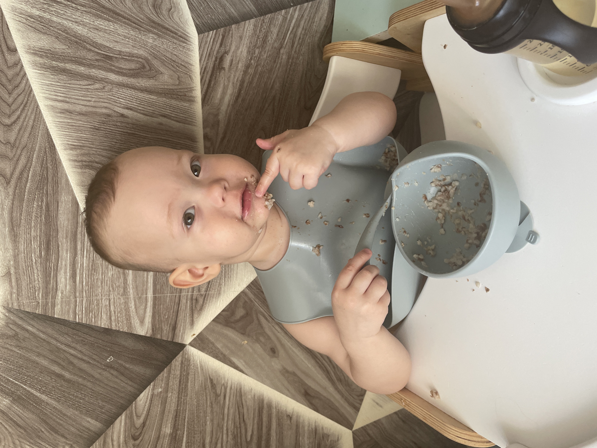 A baby is seated in a high chair, wearing a gray bib, and appears to be eating. The baby has food around their mouth and on the bib, and is holding their finger to their mouth. A gray bowl, partially filled with food, is on the high chair tray, alongside a bottle. The surroundings appear to be indoors, with a wood-patterned floor and a white high chair tray. The location is the city of Perm, Russia.