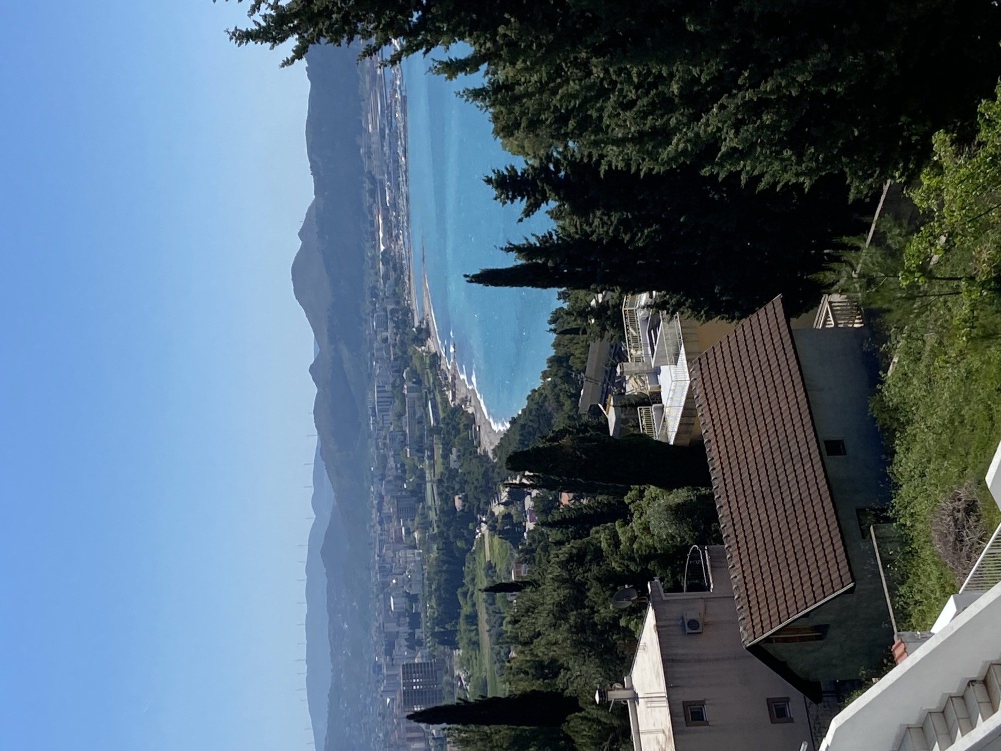 The image depicts a coastal cityscape, likely Šušanj, Montenegro, from an elevated perspective. A range of mountains is visible in the background, extending along the left side of the frame. A shoreline with a beach runs parallel to the mountains. Buildings and trees populate the area between the mountains and the coast. In the foreground, a portion of a residential building with a brown tiled roof is present. Trees are scattered around the building. The sky is clear and blue.