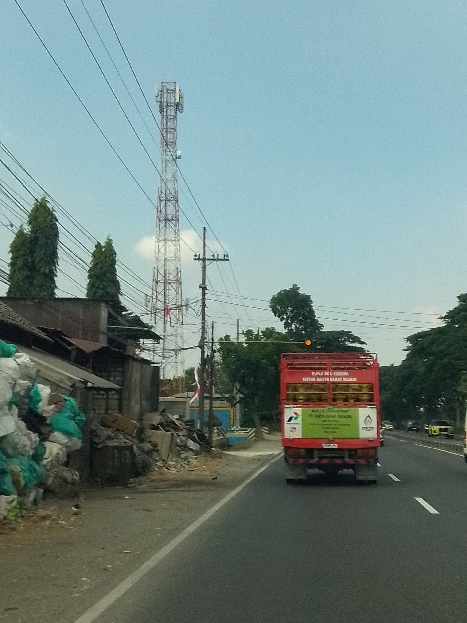 The image shows a roadside scene in Kesambi, Indonesia, on a clear day. A tall, white telecommunications tower dominates the background, with multiple wires crisscrossing the sky. In the mid-ground, a red truck is visible, carrying cylinders on its open bed. The truck has text and logos on its rear, suggesting it belongs to a company named "PT. KARYA USAHA PERSADA". To the left of the truck, there is a cluttered area with what appears to be a building or structure with a rusted roof, surrounded by piles of green and white bags. A road runs diagonally through the foreground, with a white lane marker visible. Trees and vegetation are scattered throughout the scene, with more dense foliage visible in the distance. The overall impression is of an industrial or semi-urban environment.