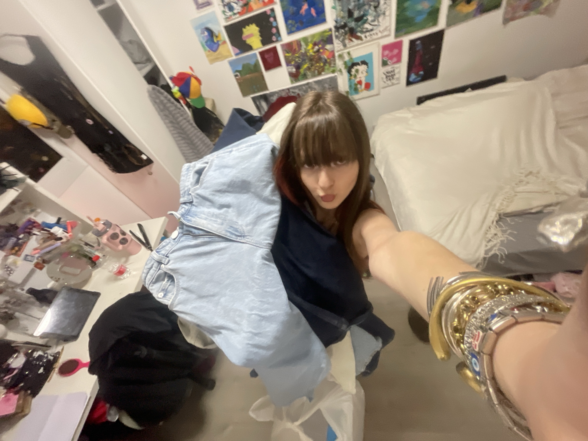 The image depicts a woman with brown hair and bangs, taking a selfie indoors. The background includes a white bed with a white comforter, a wall adorned with various colorful drawings and artwork, and a white cabinet. Clothes, including blue jeans and dark clothing, are piled in front of the woman. The scene shows a cluttered environment. The location is likely a private residence, possibly a bedroom, in Raanana, Israel.