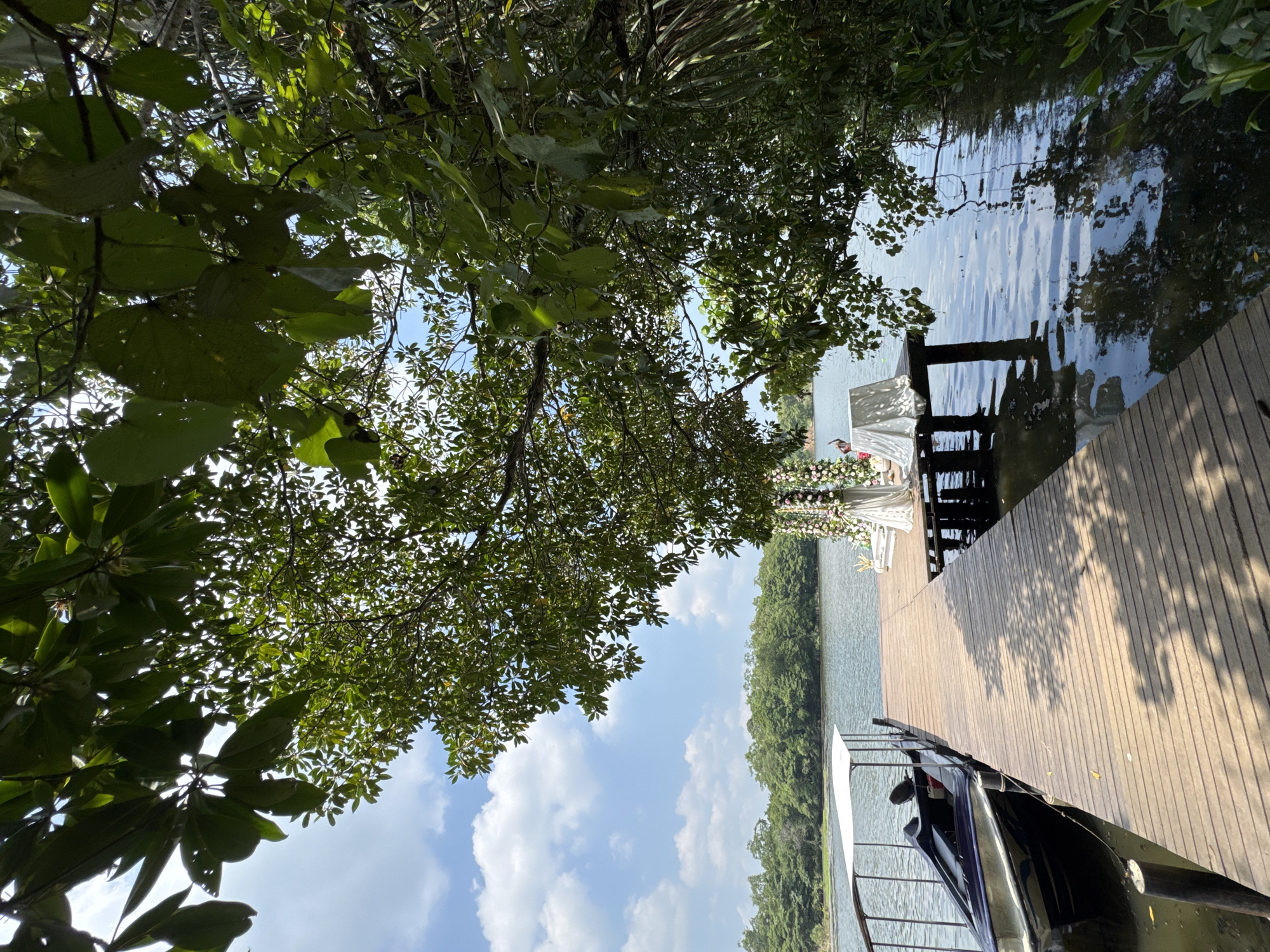 The image depicts a scene in Madapatha, Sri Lanka, featuring a wooden pier extending over a lake. The pier is lined with trees. A boat is docked on the pier in the lower right, and a wedding arch decorated with white fabric and flowers is visible at the pier's end. The water reflects the sky and trees.
