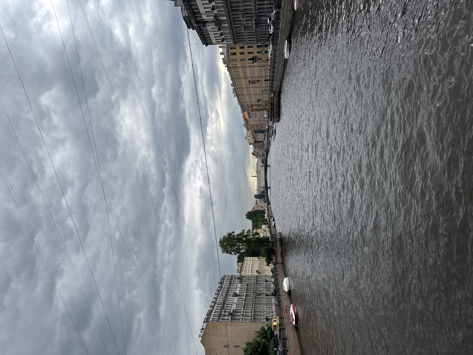 This is a landscape, eye-level, medium shot, captured on a cloudy day, of a canal in Saint Petersburg, Russia. The canal occupies the majority of the frame, its dark water rippling under the overcast sky. On the left bank, a row of historic apartment buildings with ornate facades lines the waterway. Several moored boats, including a red and white speed boat, are visible along the embankment. A solitary green tree stands near the water's edge. The right bank features a continuous line of similarly styled apartment buildings, stretching towards a distant bridge. A few cars are parked along the street behind the embankment. Overhead, a network of power lines crisscrosses the sky, partially obscuring the dense, grey clouds.