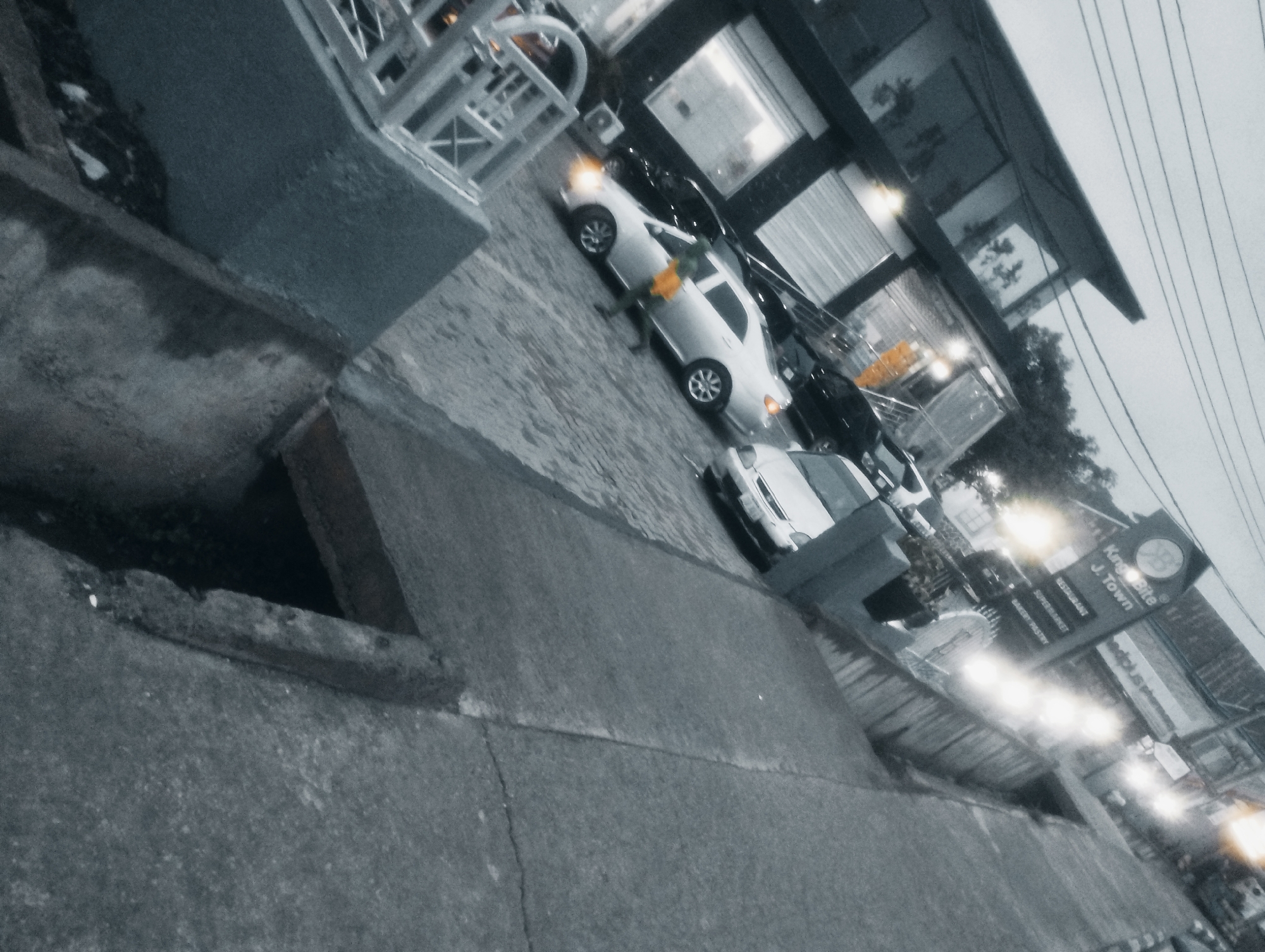 The image depicts a street scene in Jos, Nigeria, likely in the evening given the artificial lighting. In the foreground, a gray concrete barrier and curb are visible, with a dark, recessed area that may be a drainage channel or ditch. The midground features a paved street or parking area, with several cars parked and on display. A white sedan is prominent, with its headlights on, suggesting it might be for sale or recently used. Another dark-colored vehicle is parked behind it. To the right, a building with large windows and awnings is visible, housing businesses. A prominent sign reads "King's Bite J. Town" and lists services like "RESTAURANT," "SUPERMARKET," and "BAKERY PASTRY." The overall atmosphere is that of a commercial street, possibly a car lot or a strip of shops. The sky is overcast and a power line is visible crossing the upper right portion of the frame. The image is slightly tilted, giving a dynamic perspective of the scene.