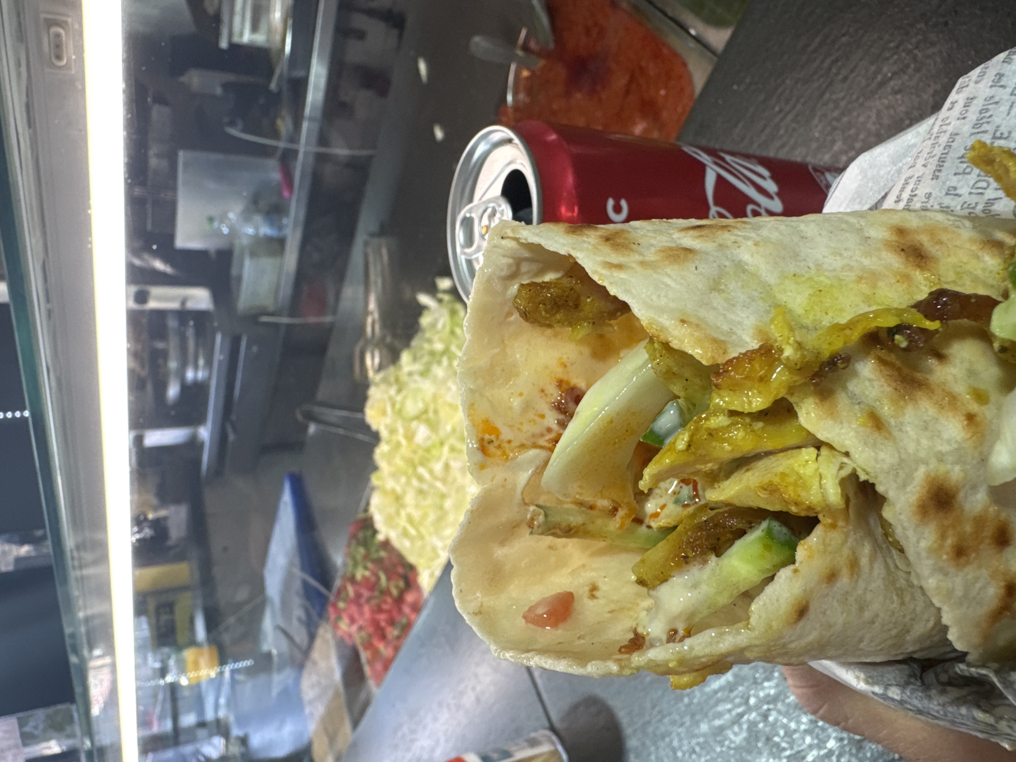 A shawarma wrap is the main subject, filled with meat, white sauce, pickles, and other ingredients. A can of Coca-Cola is partially visible in the background. The shawarma wrap is likely located in Tel Aviv, Israel, based on the commonality of shawarma in the city. The wrap is held up to a deli case with various toppings.