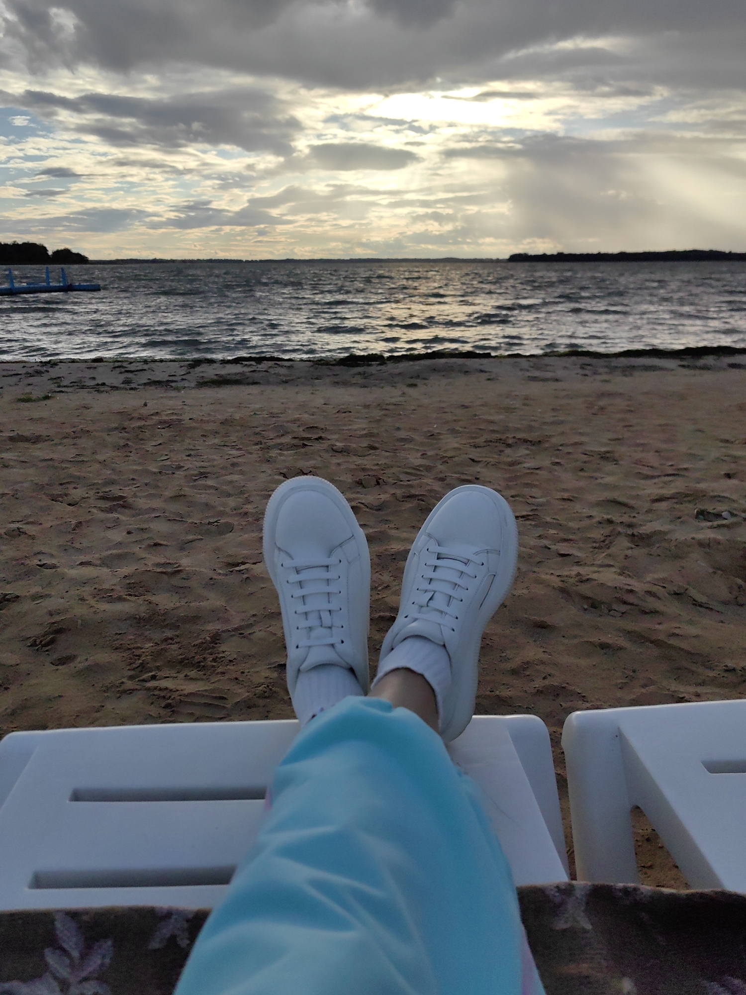 A first-person perspective shot shows a person's feet, clad in white sneakers and blue pants, resting on a white lounge chair. The background consists of a sandy beach leading to a body of water under a cloudy sky. A faint outline of land is visible on the horizon. The scene is likely located at Lake Myadel, a popular resort area in Myadzyel, Belarus, known for its beaches and recreational opportunities.