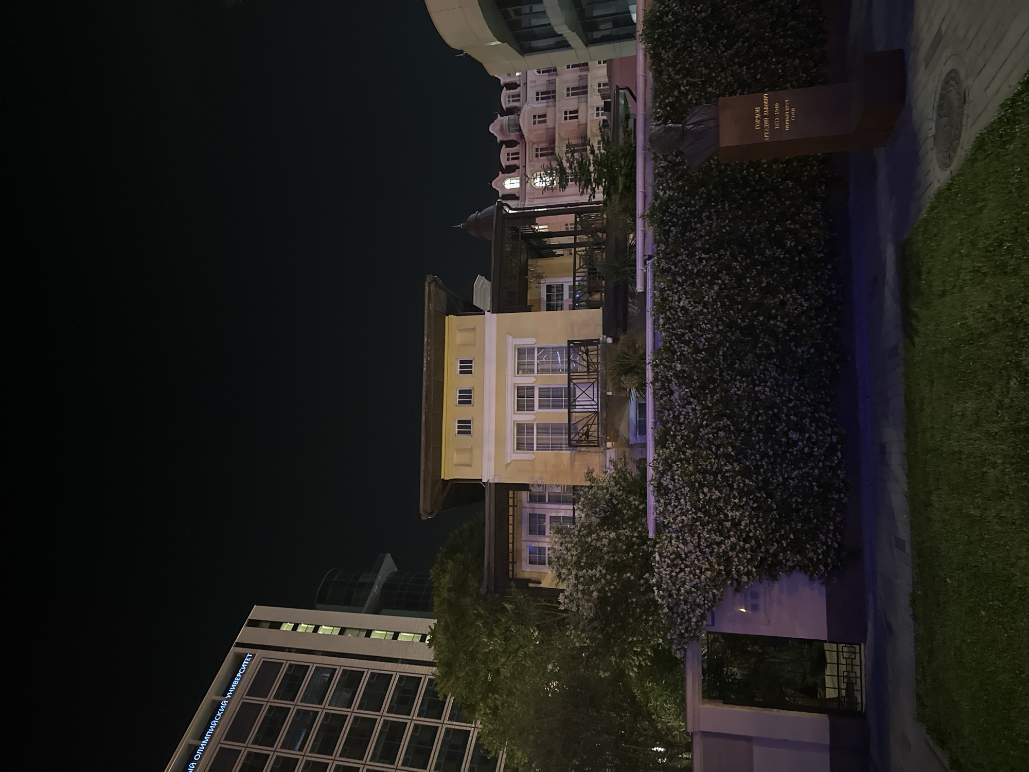 In Sochi, Russia, at night, a yellow villa with a dark wooden porch is visible in the foreground. The villa has white-framed windows and a balcony. A dense green hedge, covered in white flowers, runs along the front of the property, with a black metal gate on the left side. Behind the villa, a modern glass-fronted building with the illuminated text "СОЧИНСКИЙ ОЛИМПИЙСКИЙ УНИВЕРСИТЕТ" (Sochi Olympic University) is present, along with other buildings of varying architectural styles, including a light-colored building with arched windows and a domed roof. A dark-colored statue of a bust on a pedestal stands to the right of the hedge, with text in Cyrillic. The scene is lit by artificial lights.
