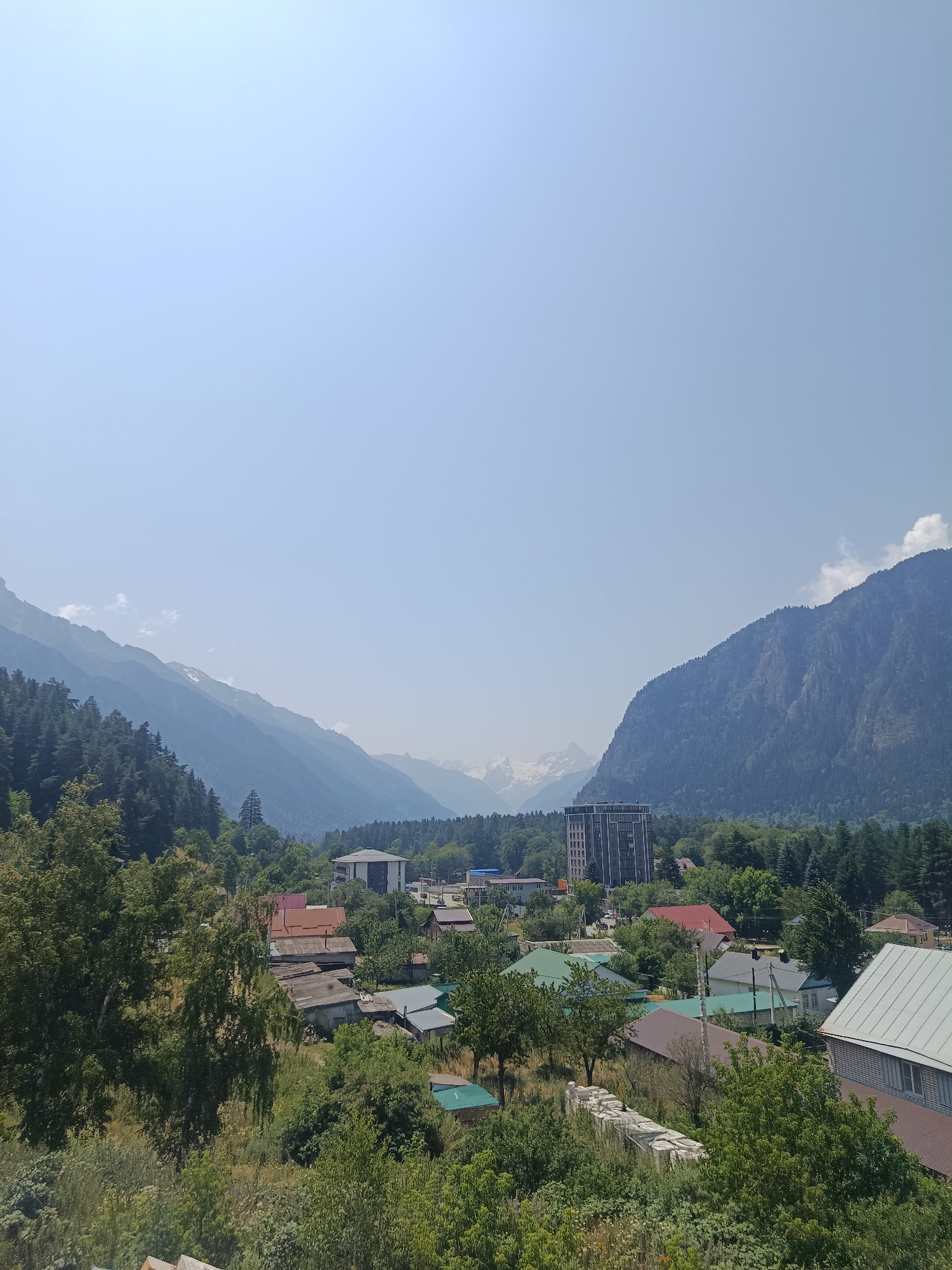 The image depicts a scenic view of the city of Teberda, Russia, nestled within a valley surrounded by majestic mountains. In the foreground, dense green foliage and trees partially obscure a cluster of residential buildings with various roofing materials, including red, gray, and green. A few of these houses appear to be older, constructed with what looks like aged wood and stone, while others are more modern. A tall, dark-colored building, likely an apartment complex or hotel, stands out in the mid-ground, contrasting with the surrounding low-rise structures and natural landscape. In the background, imposing mountains rise sharply, their slopes covered in dark green coniferous forests. The higher peaks are snow-capped, indicating significant elevation. The sky is clear and bright blue, suggesting a sunny day. The overall scene conveys a sense of peaceful coexistence between a small town and its dramatic natural environment.