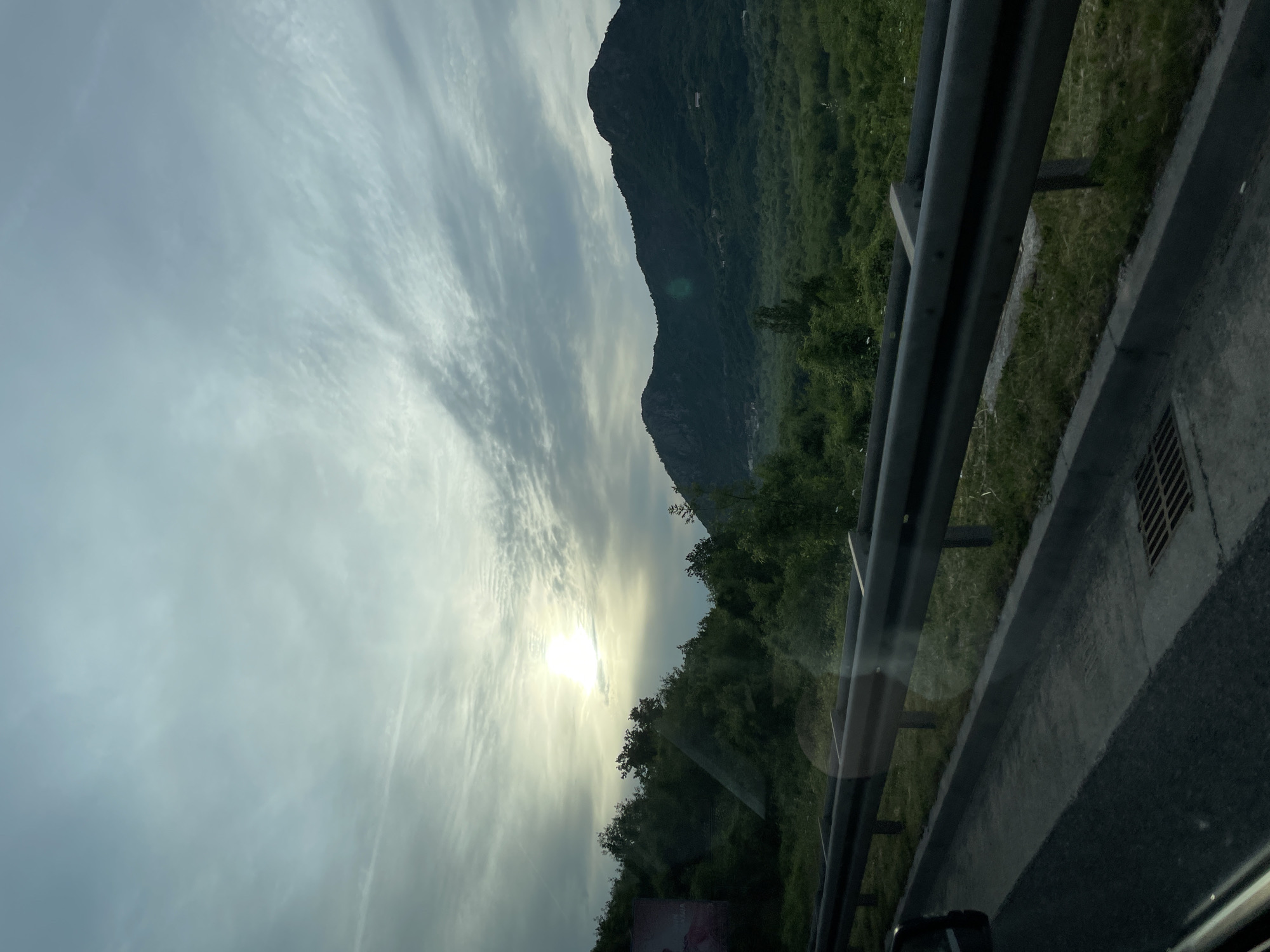 The image shows a landscape in Sotonići, Montenegro, likely taken from a moving vehicle. The view is oriented vertically, with a cloudy sky overhead. The sun is visible, partially obscured by clouds. A mountain is present in the background, covered with greenery. A metal guardrail is in the foreground, alongside a concrete barrier and a section of asphalt road. Vegetation is visible beyond the guardrail. A drainage grate is located near the edge of the road.