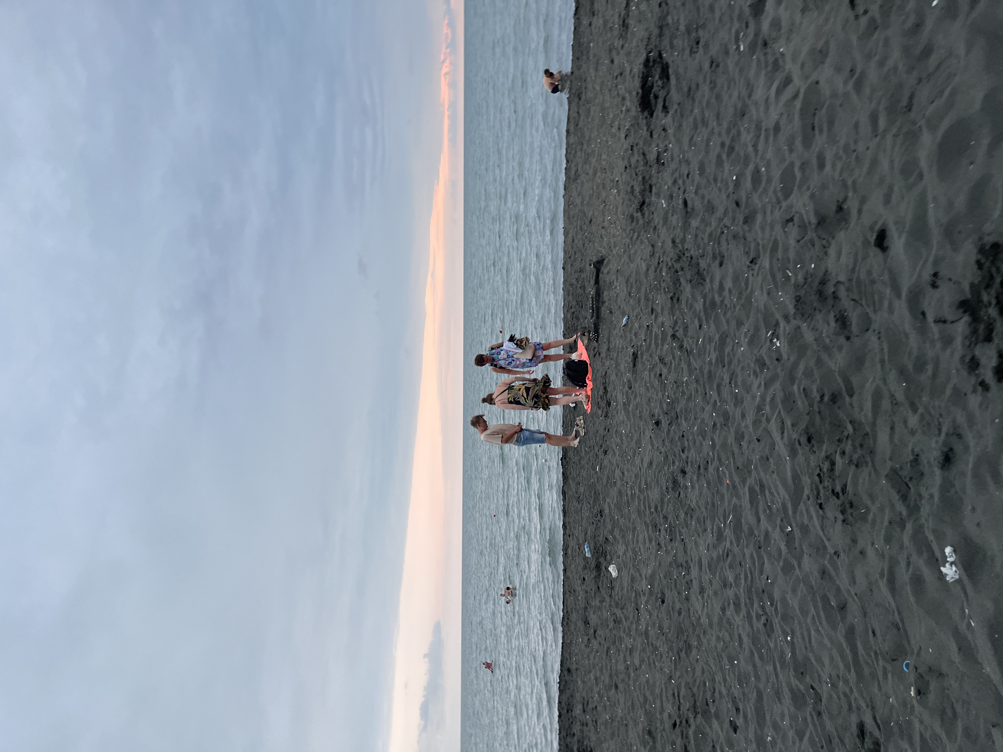 The image depicts a beach scene in Ureki, Georgia, under a cloudy sky with a soft pink and orange glow on the horizon. The beach is covered in dark, fine-grained sand, with some scattered debris visible. In the foreground, three people stand near the water's edge. Two individuals, appearing to be a couple, are looking down at something on the sand. The third person, a woman, is facing away from the camera. Nearby, a bright orange bag lies on the sand, along with a pair of sandals and a black item that could be a towel or a bag.
Further out in the calm, grey sea, several people are swimming. One person is visible further out in the water, seemingly engaged in an activity like surfing or paddleboarding, indicated by a red object they are holding. Another person is crouched at the water's edge on the right side of the frame. The overall atmosphere is serene, with a hint of human activity on a tranquil beach.