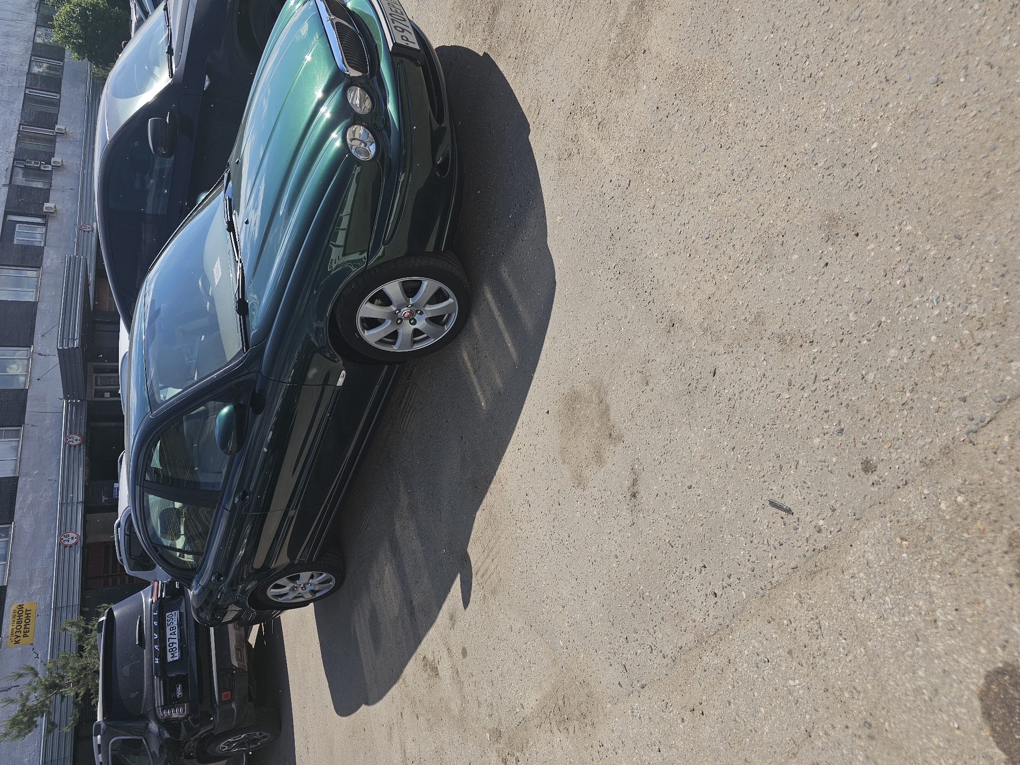 An outdoor photograph, rotated 90 degrees clockwise from its natural orientation, depicts a street scene with several parked cars under bright sunlight. The prominent subject is a dark green Jaguar X-Type sedan, parked on an asphalt surface. Its front is angled towards the top right of the frame, showcasing its circular headlights and chrome grille. The car's front license plate, "P 970 EP 190," is clearly visible, indicating a Russian registration (Moscow region).
Behind the Jaguar, further to the left, a dark SUV, identifiable as a Haval, is also parked. Its rear is visible, showing the license plate "M 897 AB 550," another Russian registration. A third, darker car is partially visible further back.
The ground is light-colored asphalt, casting strong, defined shadows from the cars, suggesting bright sunshine and either mid-morning or afternoon. Scattered debris and small stones are visible on the road surface.
In the background, a multi-story, grey building with numerous rectangular windows and visible external air conditioning units lines the street. A yellow sign is attached to the building's facade on the far left, displaying the text "КУЗОВНОЙ РЕМОНТ" (BODY REPAIR), along with what appears to be a phone number or registration: "(C)109114-193." Below this sign, a circular red and white sign with the number "3.6" is also visible, likely indicating a height restriction. Green foliage from a tree is seen near the top left corner. The overall scene suggests an urban setting in Russia.