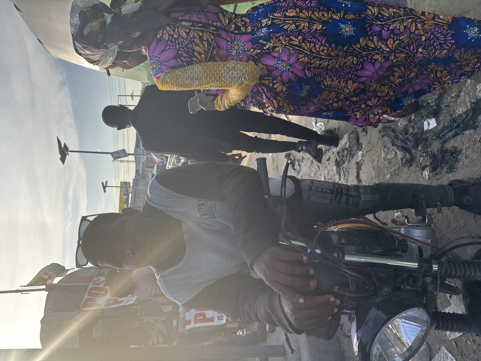 The image depicts an outdoor, urban commercial scene in Lagos, Nigeria, likely in the late afternoon or early morning given the strong sun glare.
In the left foreground, a dark-skinned man, wearing a grey shirt with dark long sleeves featuring "BROOKLYN" text, and sunglasses perched on his head, is seated on a black motorcycle. His hands are clasped on the handlebars, and he is looking directly towards the camera.
In the midground to the right, a dark-skinned woman is viewed from behind, wearing a brightly patterned dress with purple, blue, and orange floral motifs, and a distinct yellow textured sleeve. She holds a light-colored smartphone in her left hand and has a patterned headscarf covering her head. Slightly ahead of her and to the left, also viewed from behind, is a dark-skinned man dressed in a dark suit jacket and trousers, walking away from the camera.
The background on the left features a white sign with prominent red "POS" lettering, indicating a Point of Sale service, partially obscured by sun glare. Behind this sign is a light-colored canopy or tent structure, with a flag (green and white with a central emblem) flying on a pole above it. Utility poles with solar panels are visible in the distant background. The ground is unpaved, consisting of dirt and scattered debris, including plastic waste. The strong sunlight from the upper left creates significant lens flare across the scene.