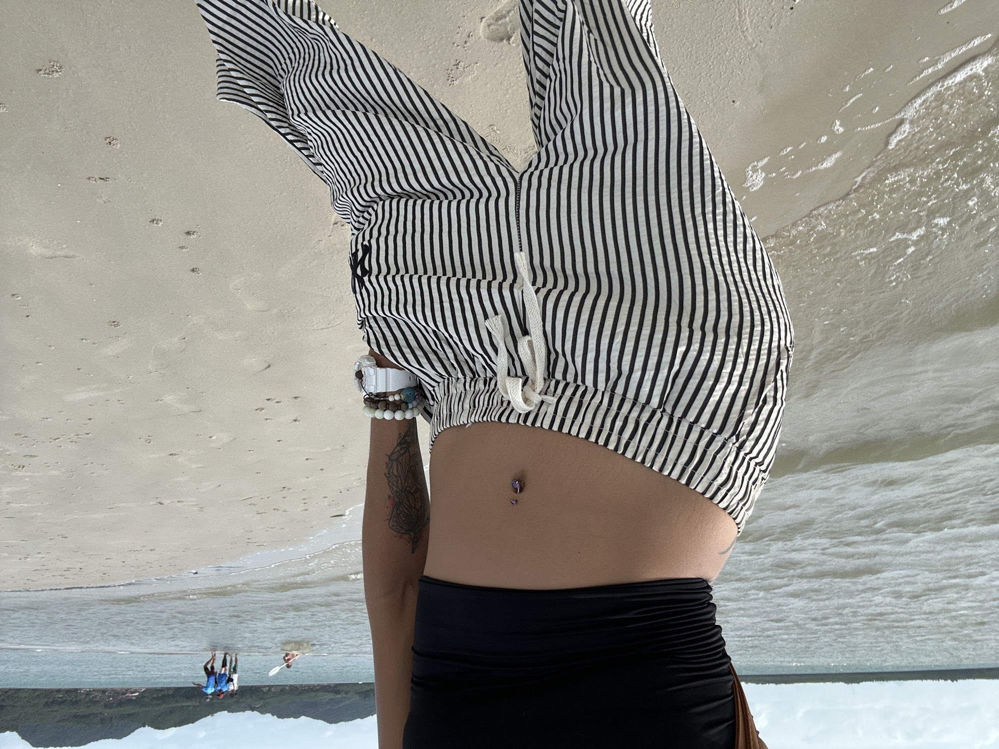 The image shows a person standing on a sandy beach with the ocean in the background. The person is wearing a black crop top and black and white striped pants. They have a belly button piercing and a tattoo on their arm. There are other people in the water in the distance, some of whom appear to be walking. The environment is a beach with sand, water, and a cloudy sky. The location is identified as Ko Yao, Thailand.