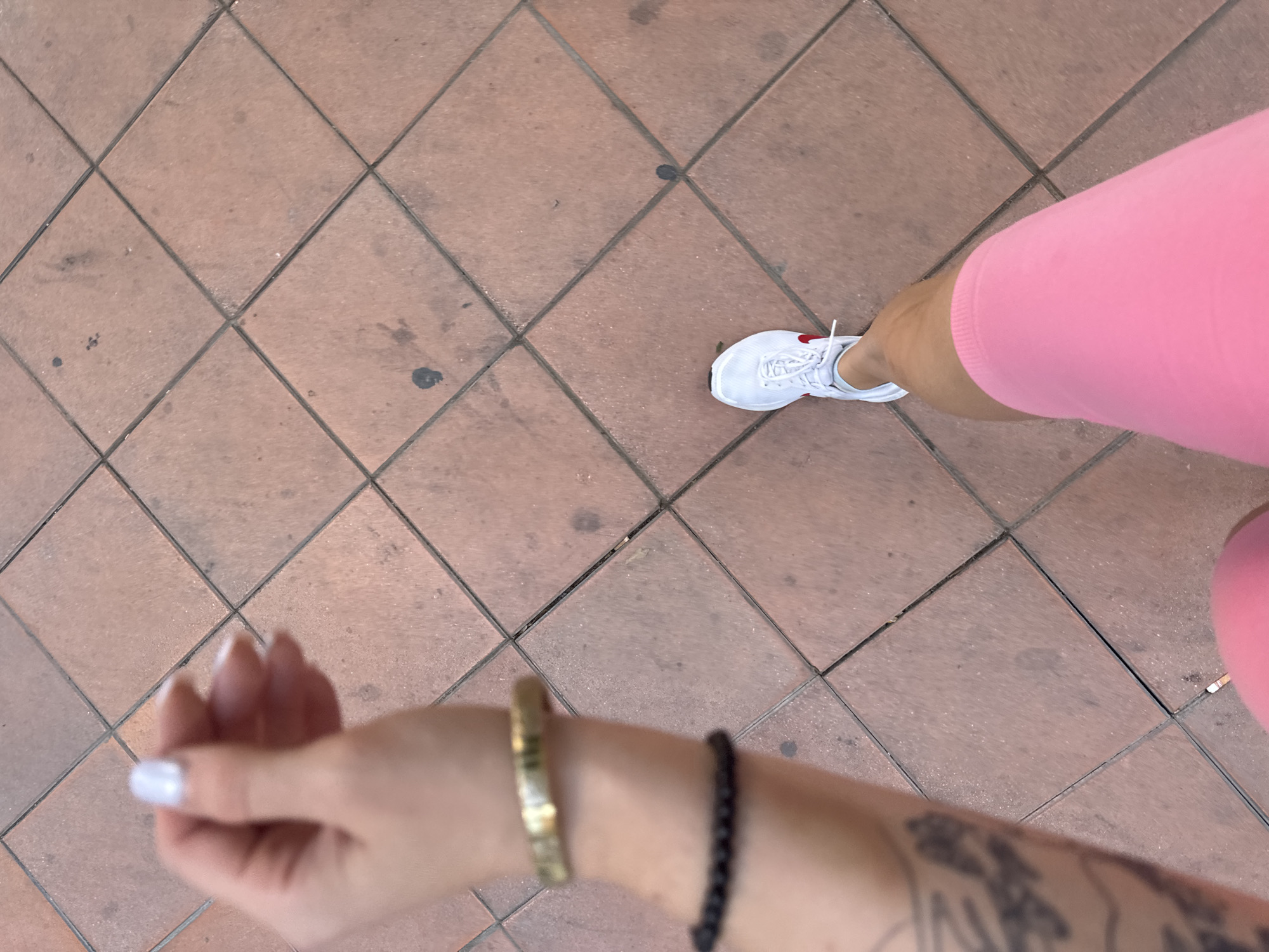The image shows a close-up, downward-facing perspective of a person walking on a tiled sidewalk. The tiles are reddish-brown and arranged in a diamond pattern, with dark grout lines. The person's left arm and hand are in the foreground, adorned with a gold-toned bracelet and a black beaded bracelet. The hand has manicured nails painted a pearlescent white. A tattoo is partially visible on the forearm. In the mid-ground, the person's right leg is visible, wearing pink biker shorts and a white athletic shoe with a red accent. The shoe is in motion, suggesting the person is walking. The scene appears to be outdoors in a city. There is no specific information within the image that would place this scene in the city of Murcia, Spain.