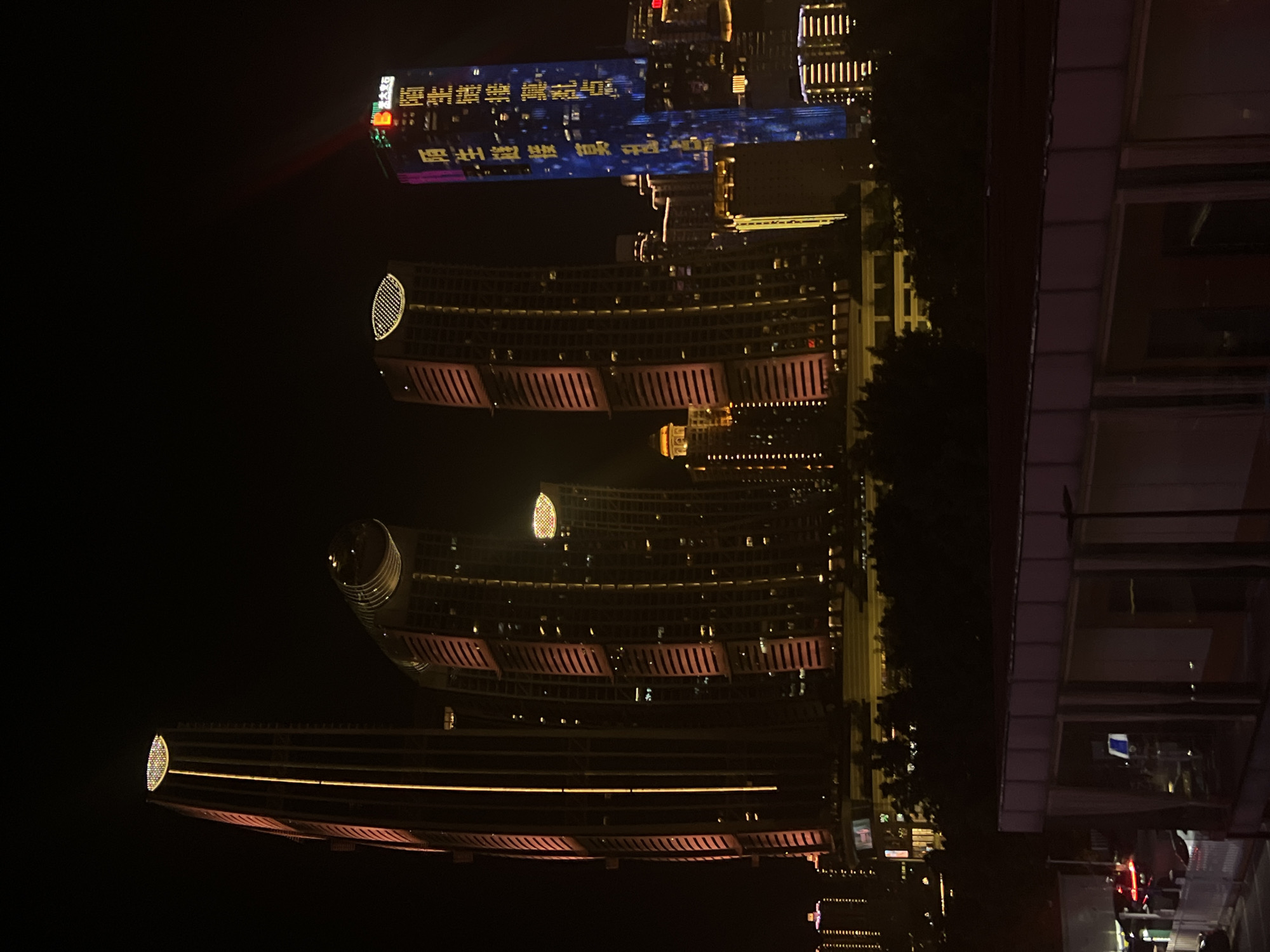 The image depicts a nighttime cityscape, likely in Jiangbei Cheng Street, China, dominated by illuminated skyscrapers. The most prominent structures are tall, modern buildings, some with curved facades, adorned with vertical lines of light in pink and orange hues. One particularly tall, slender building on the left features a light strip running its entire height and a circular, illuminated crown. To its right, a wider, curved building also displays pink lighting. Further to the right, a blue-lit skyscraper features vertical Chinese characters in yellow. In the background, other buildings are visible, some with illuminated windows and arrays of lights. A red beam of light emanates from the top of the blue-lit skyscraper. The foreground shows a dark area with trees and the top of a lower building with horizontal window-like structures and subtle lighting. The overall scene conveys a vibrant, urban environment at night, characterized by architectural illumination.